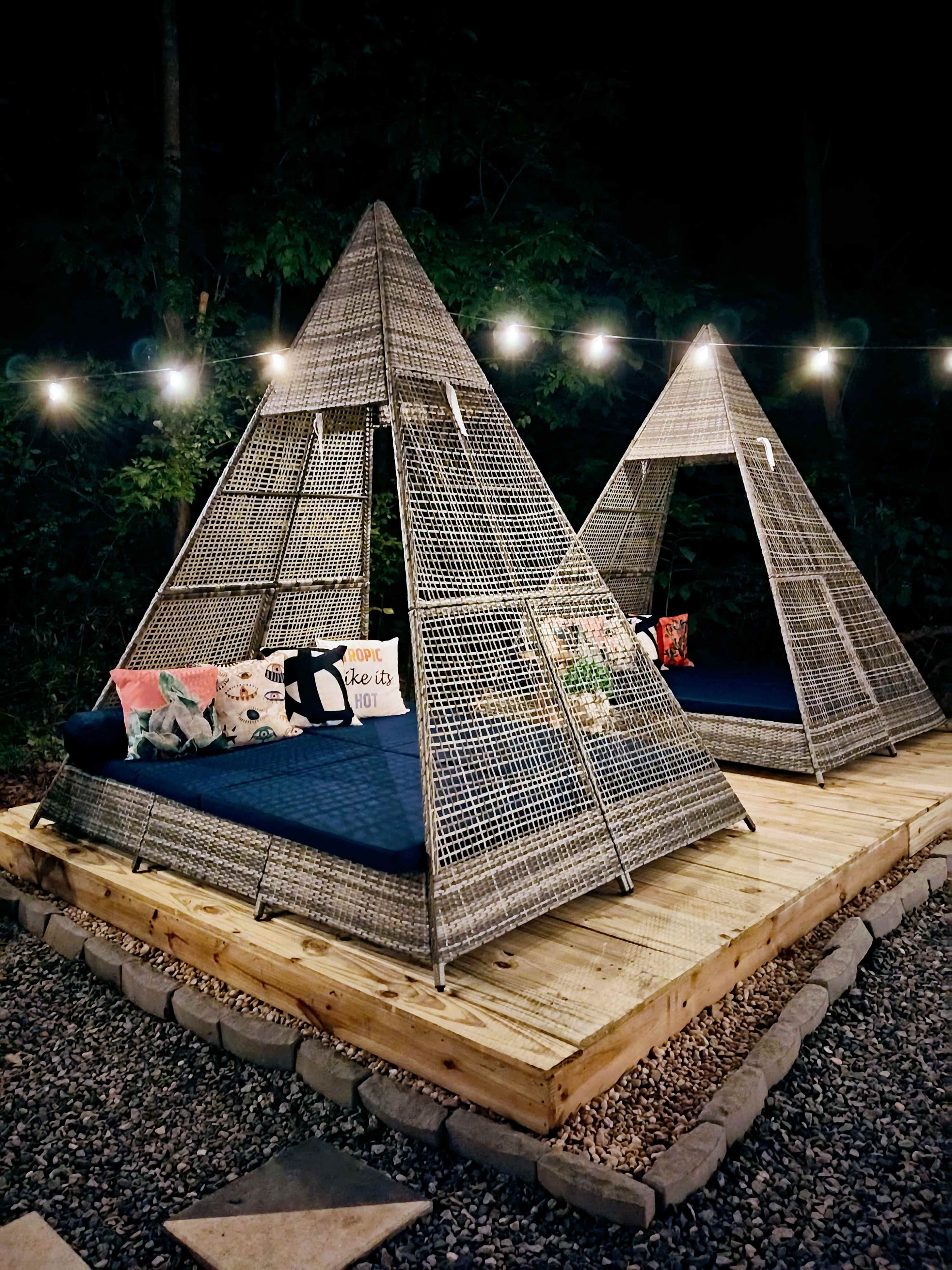 Two triangular-shaped, woven seating structures are illuminated by string lights on a wooden platform surrounded by gravel.