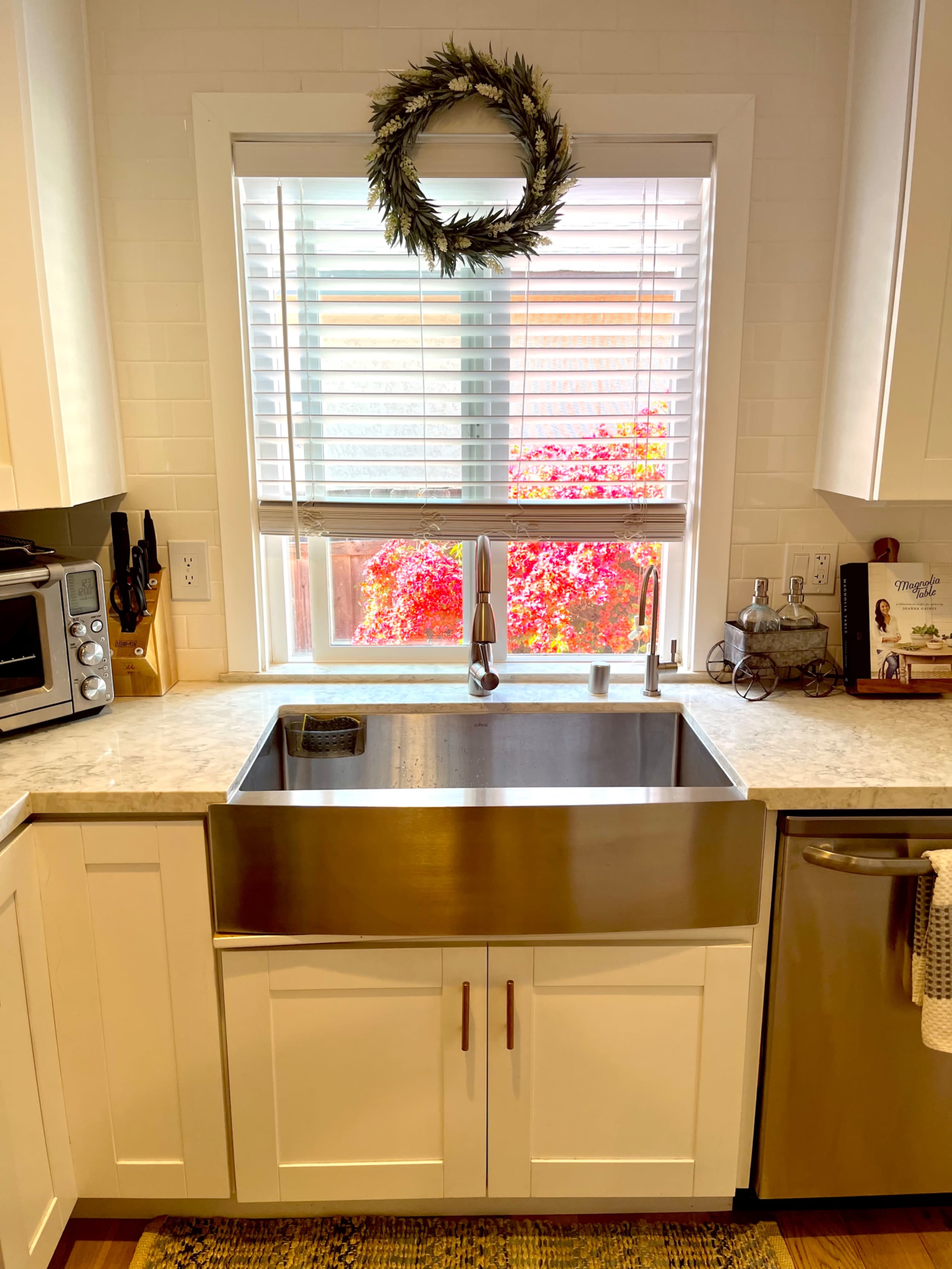 A stainless steel farmhouse sink is positioned beneath a window with blinds, overlooking a colorful outdoor garden.