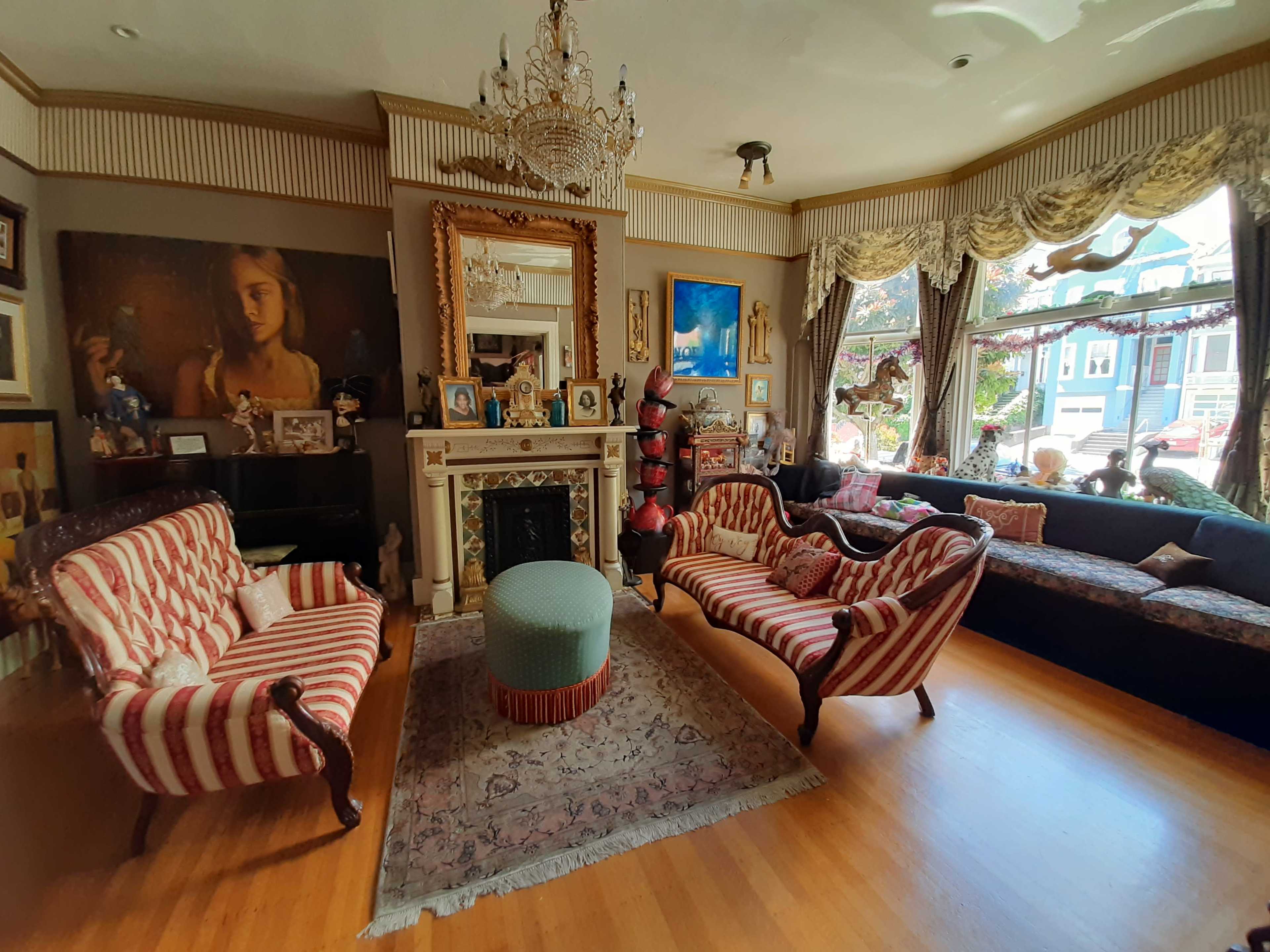The room features a mix of striped sofas and a round ottoman, with a decorative fireplace and various art pieces adorning the walls.