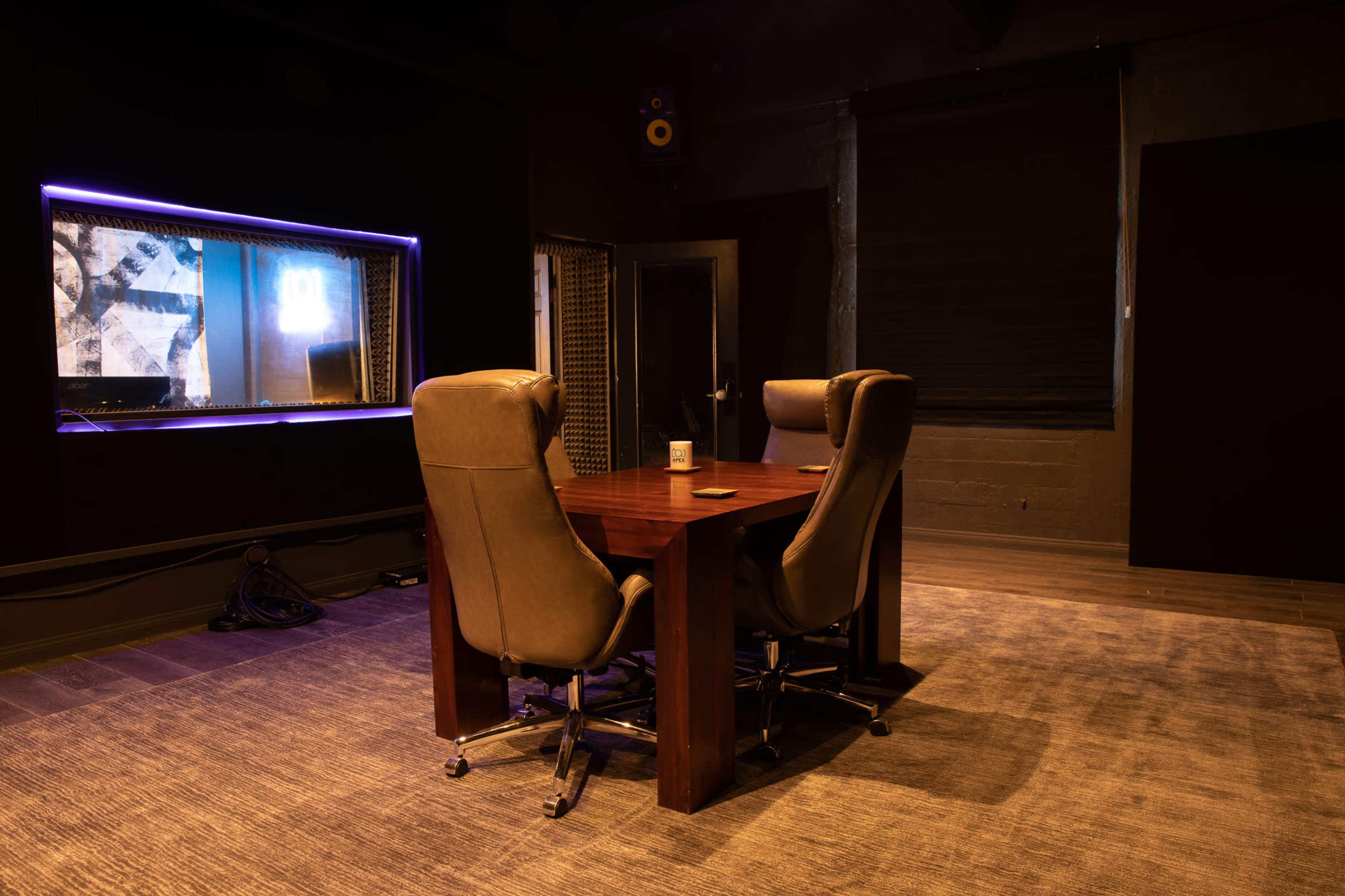 A modern conference room features a wooden table with two high-backed chairs and a large window overlooking a soundproof area.