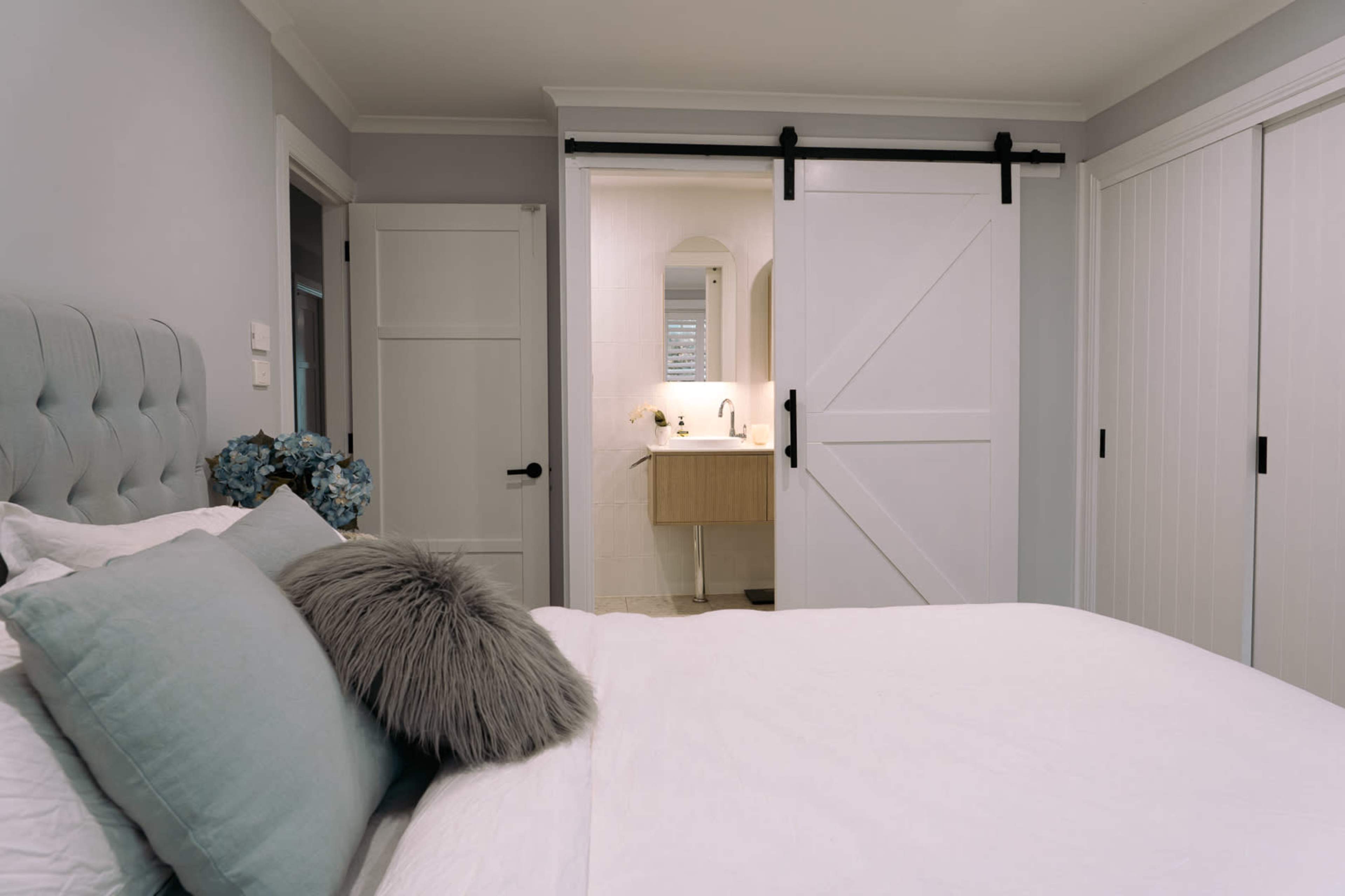 The image shows a modern bedroom with a sliding barn door leading to a bathroom area, featuring a bed with pillows and a decorative throw.