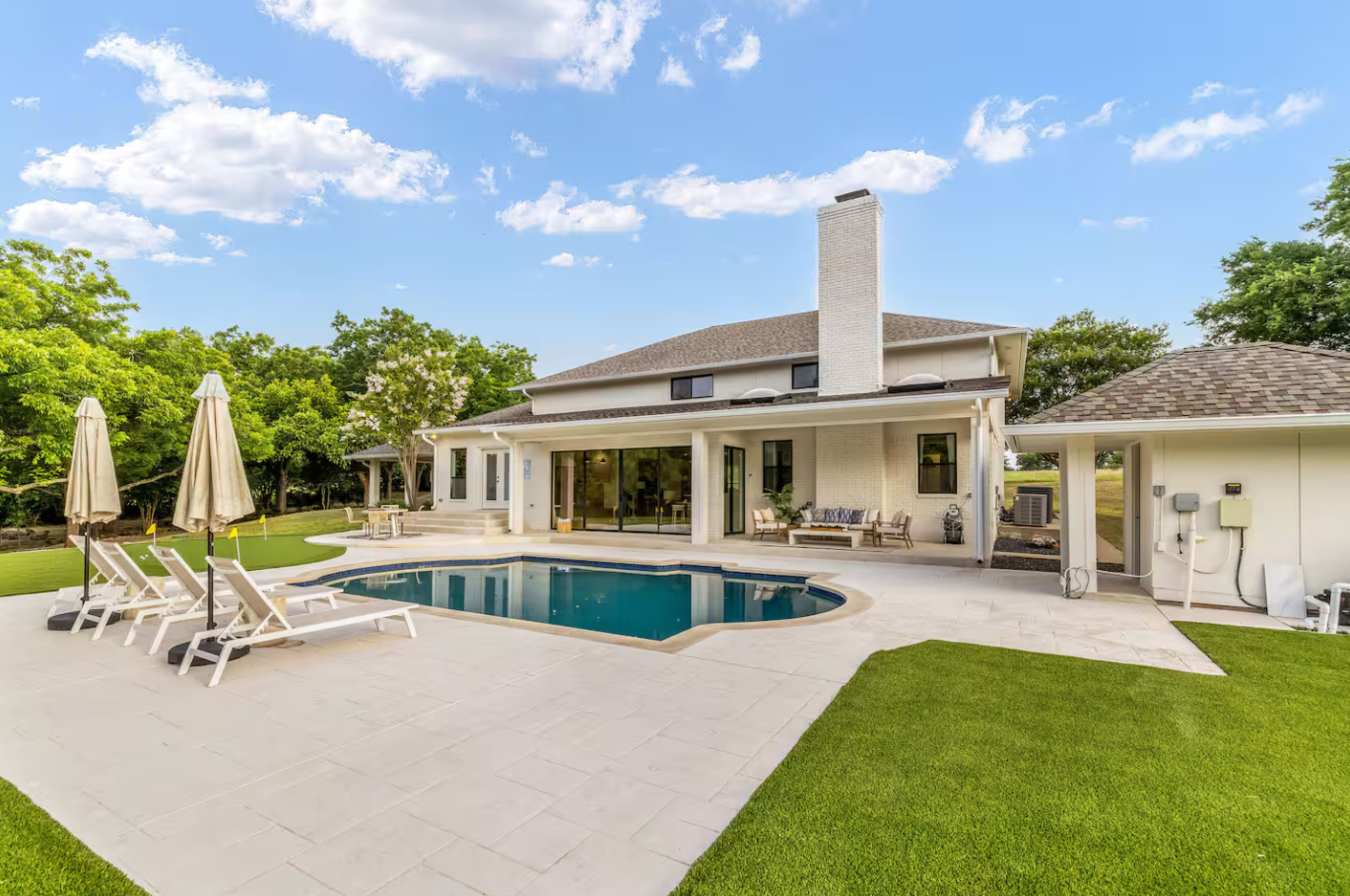 The image shows a modern house with a swimming pool surrounded by lounge chairs, set in a landscaped yard featuring green grass and trees.