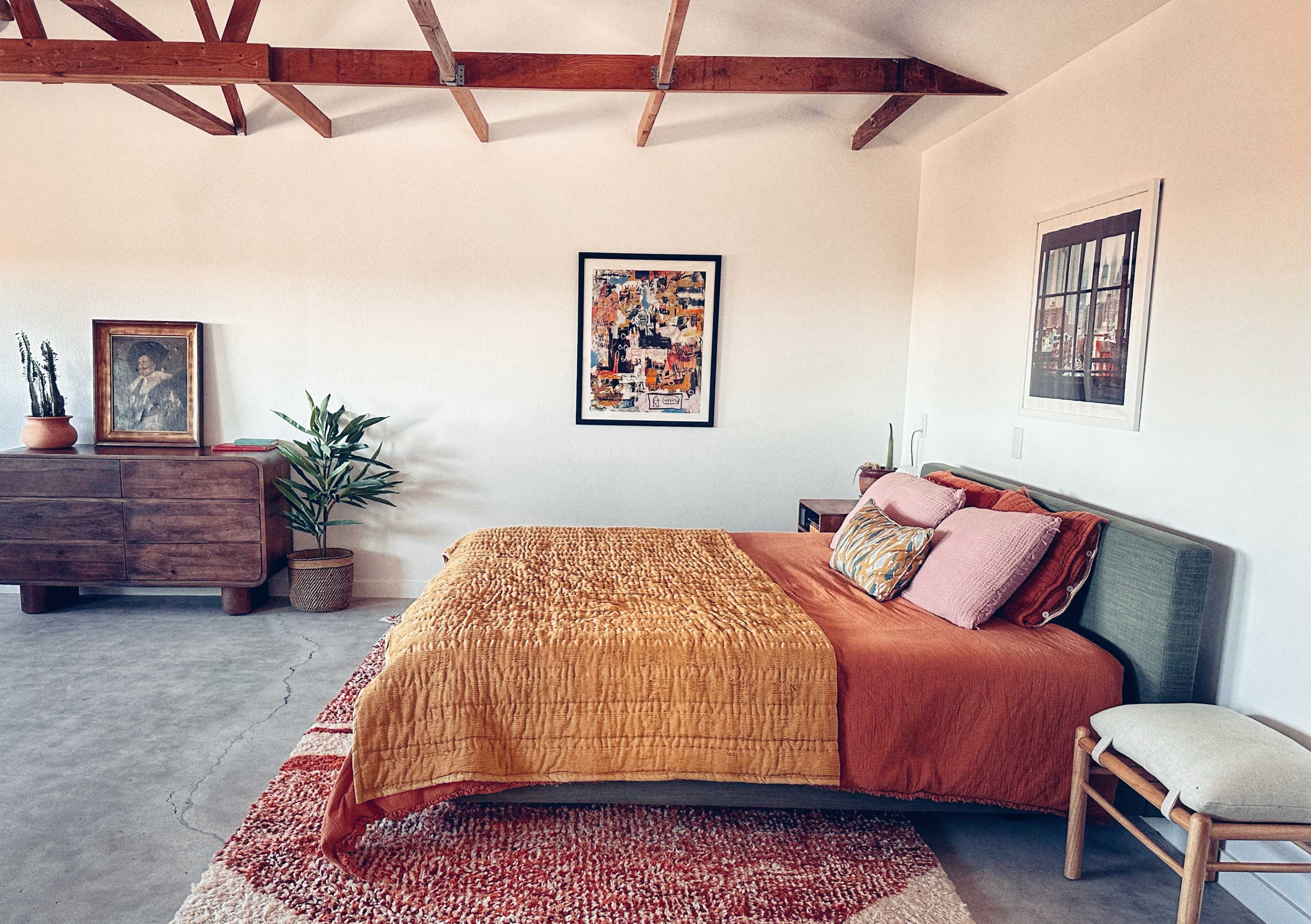 A minimalist bedroom features a bed with an orange and mustard quilt, a wooden dresser, a potted plant, and exposed wooden beams in the ceiling.