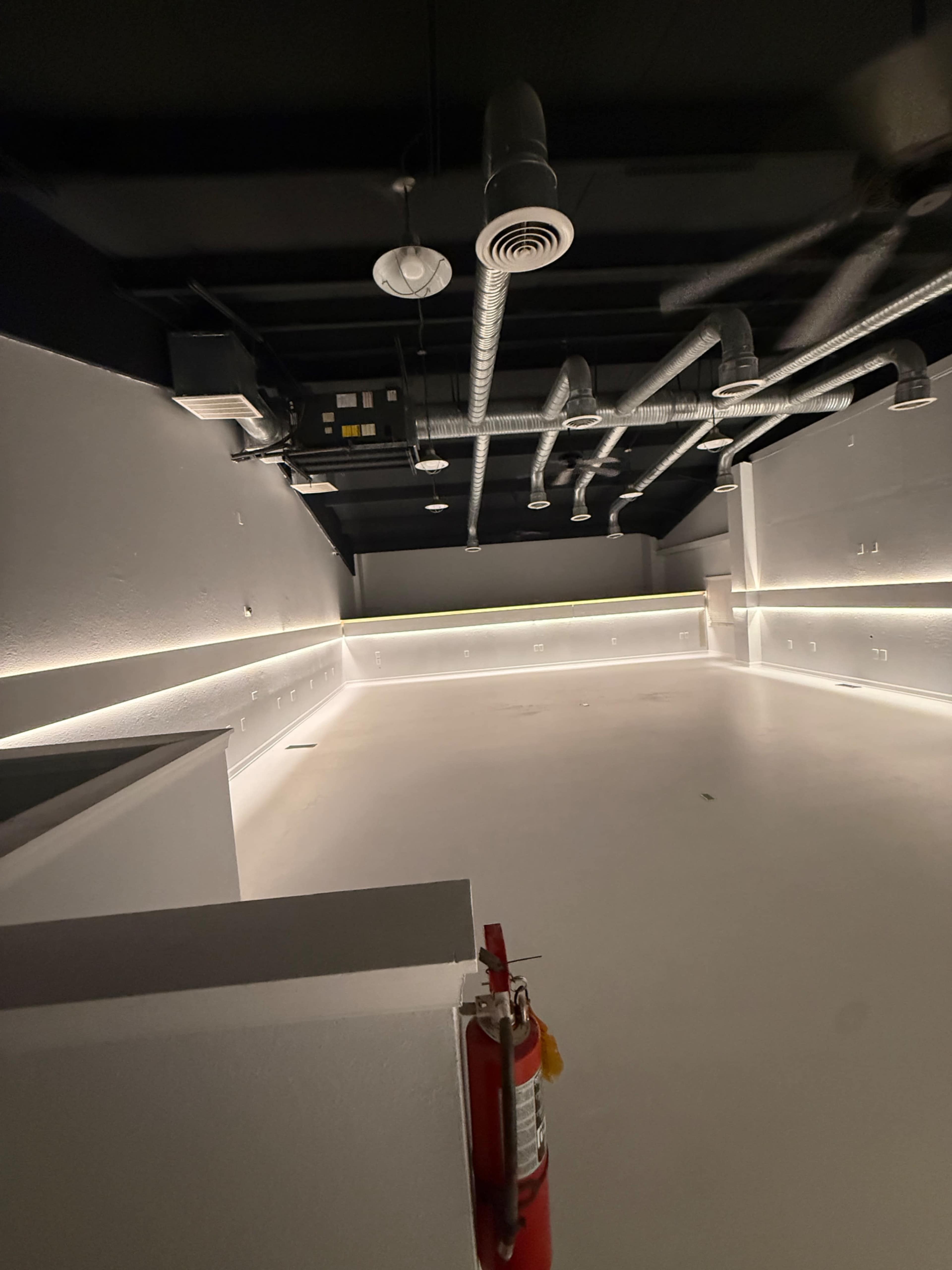 The image shows an empty room with white walls and a white floor, featuring exposed ducts and soft lighting along the perimeter.