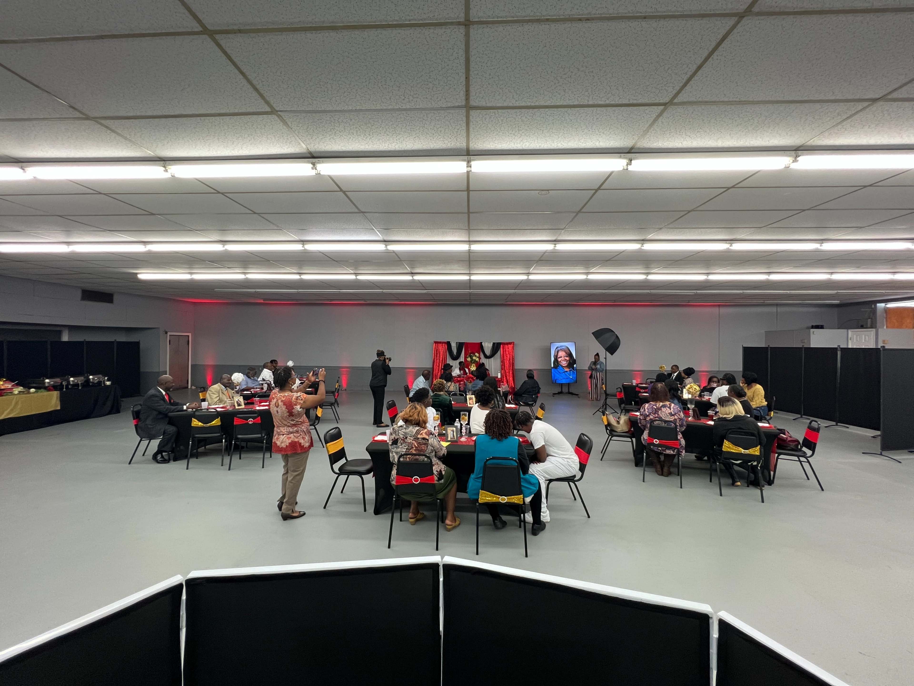 A gathering is taking place in a spacious room with tables arranged for attendees, who are engaged in various activities while a presentation occurs at the front.