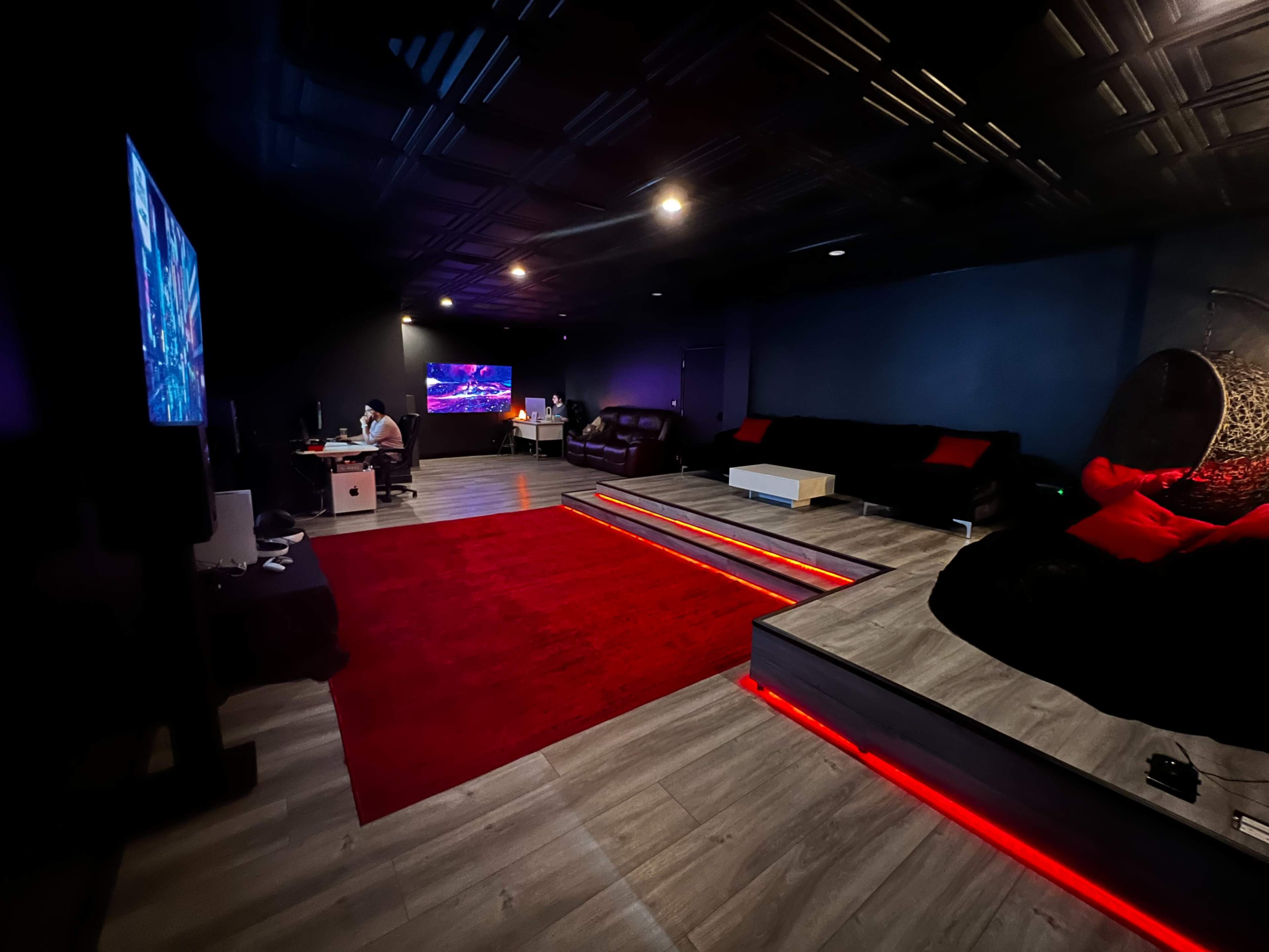A dimly lit entertainment room featuring two large screens, a red area rug, tiered seating, and cozy seating arrangements.
