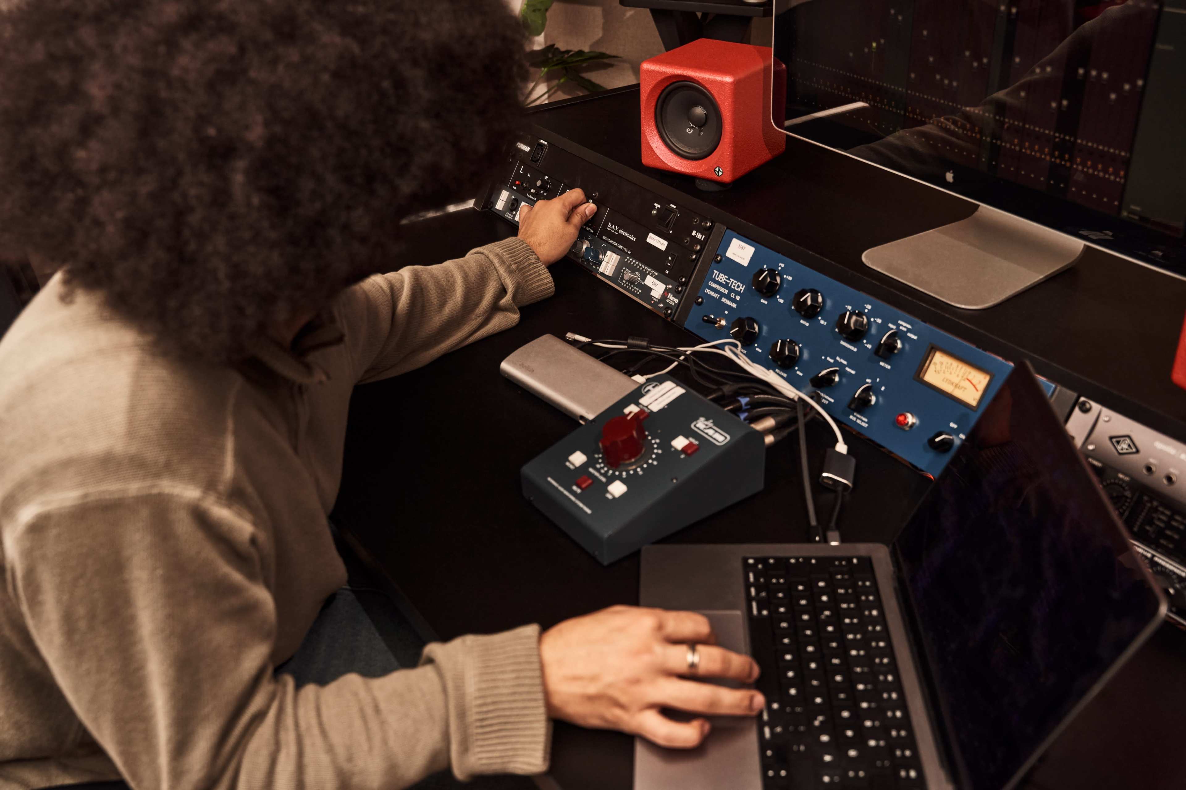 A person adjusts audio equipment on a desk with a laptop and a speaker in a music production setting.