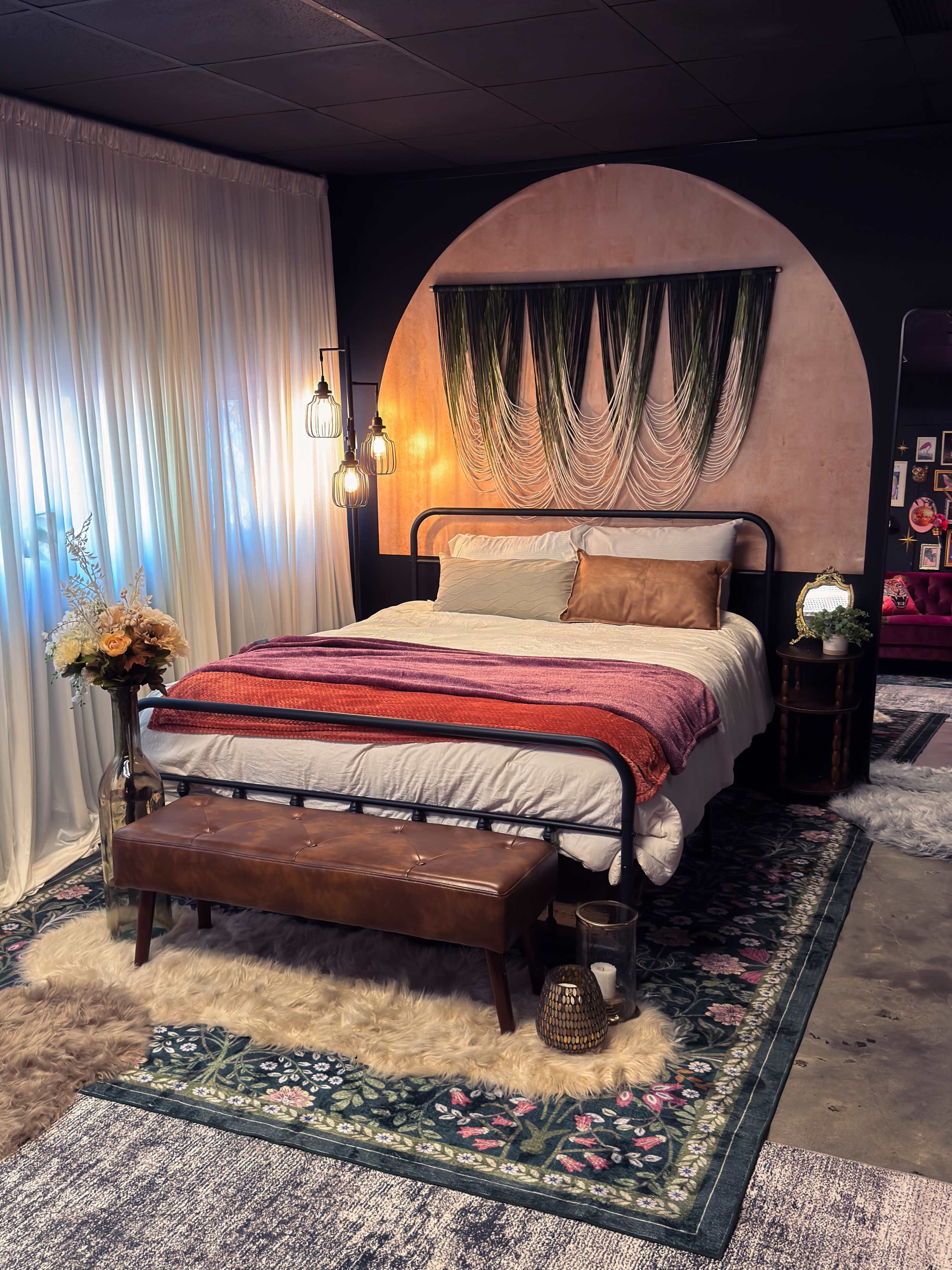The image shows a cozy bedroom featuring a metal bed frame with colorful blankets, a decorative backdrop with hanging plants, and a mix of soft rugs and decorative elements.