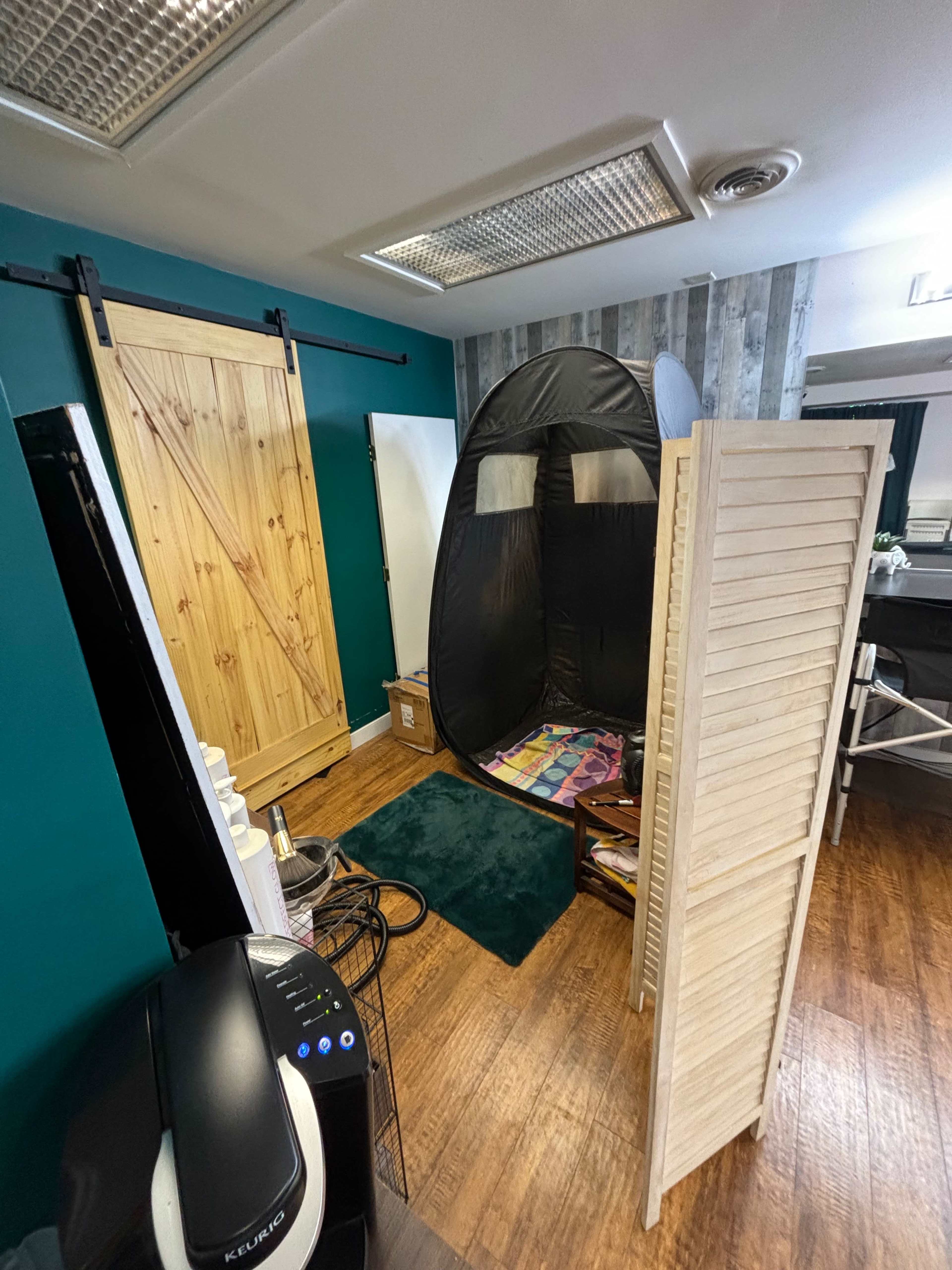 A small room featuring a wooden door, a privacy screen, a black tent-like structure, and various items on the floor.