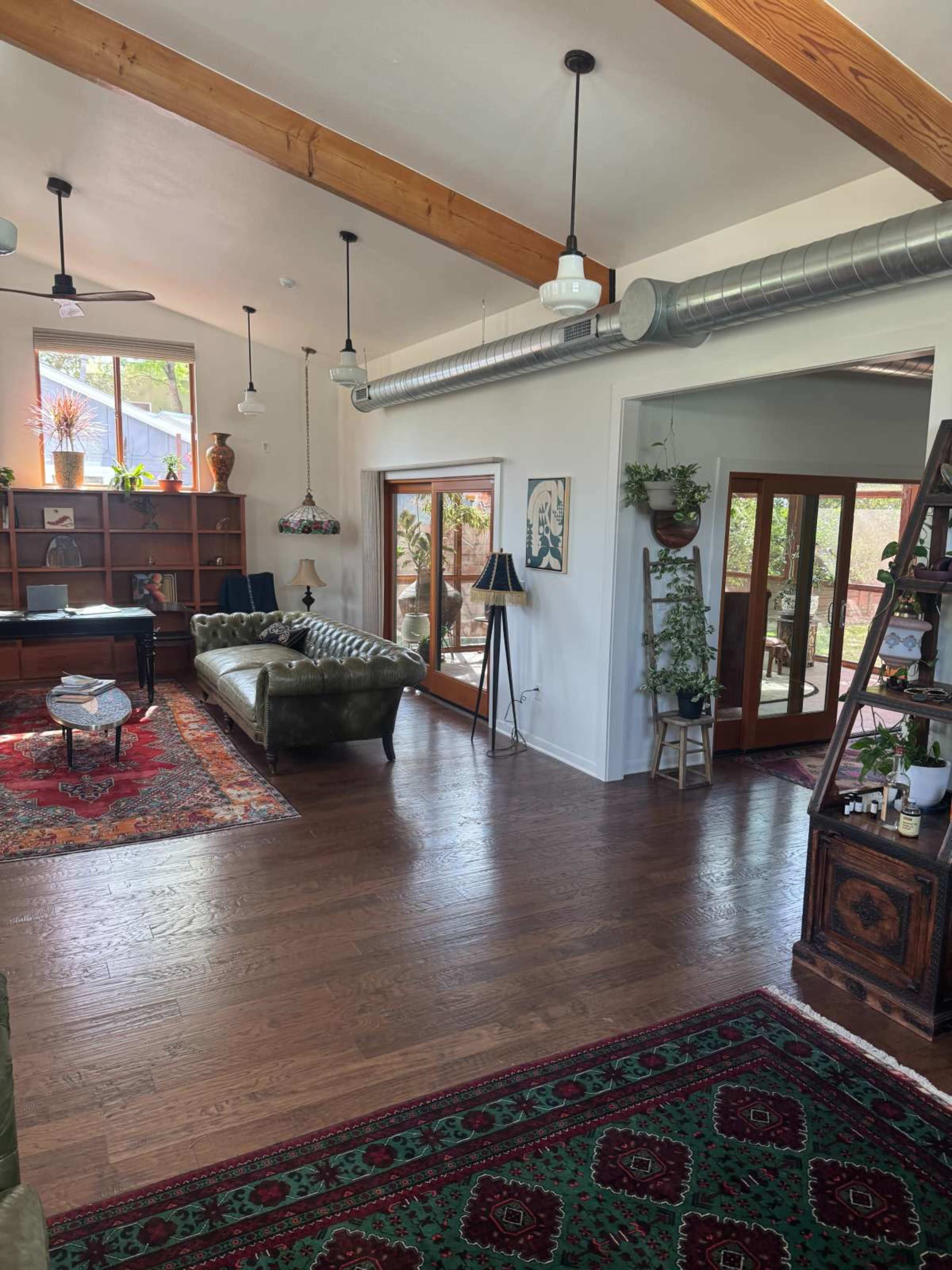 The image shows a well-lit living room with wooden beams, a piano, a green leather sofa, and large windows overlooking a patio area.