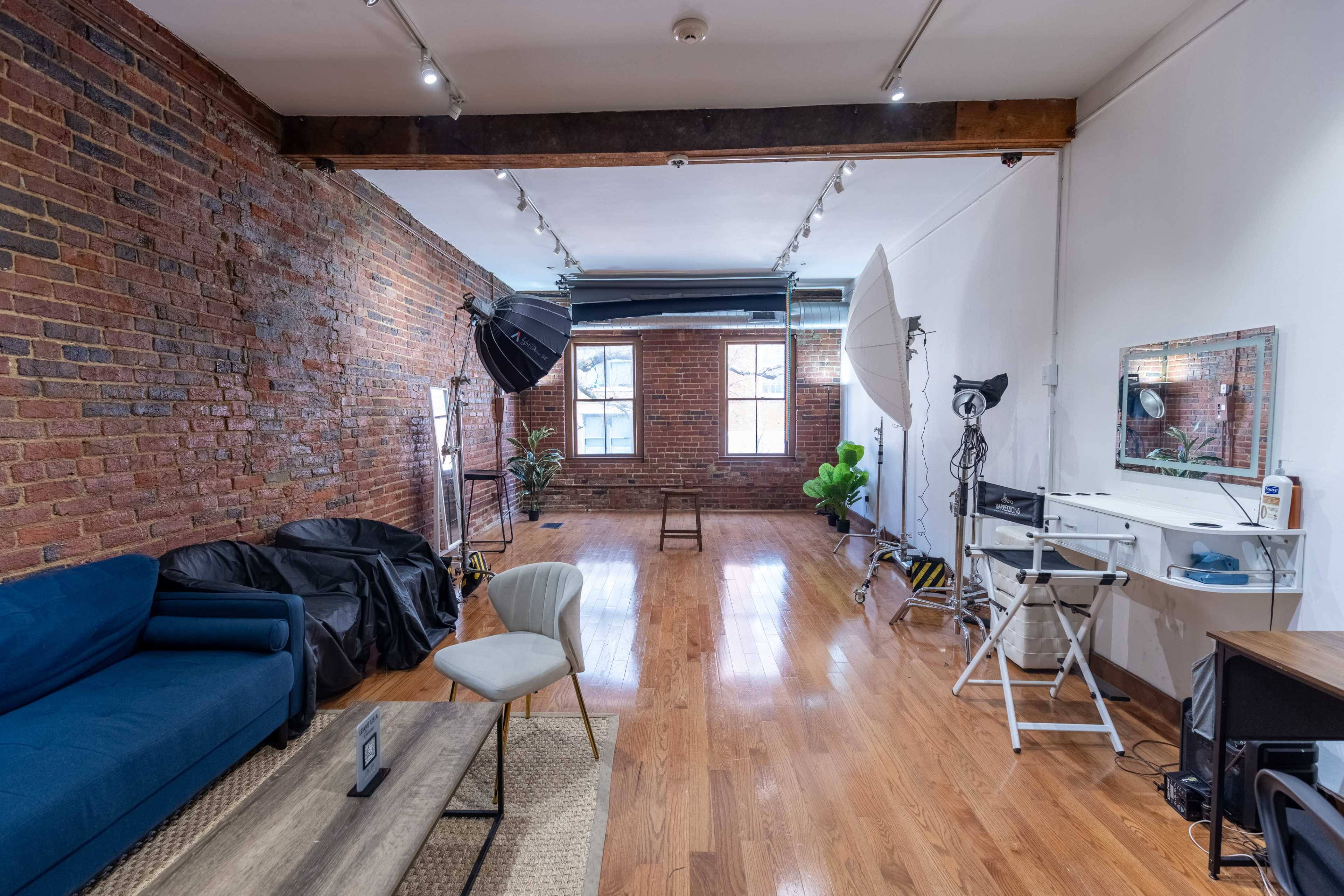 A spacious room with exposed brick walls, hardwood floors, and various photography equipment set up, including softboxes and a makeup area.