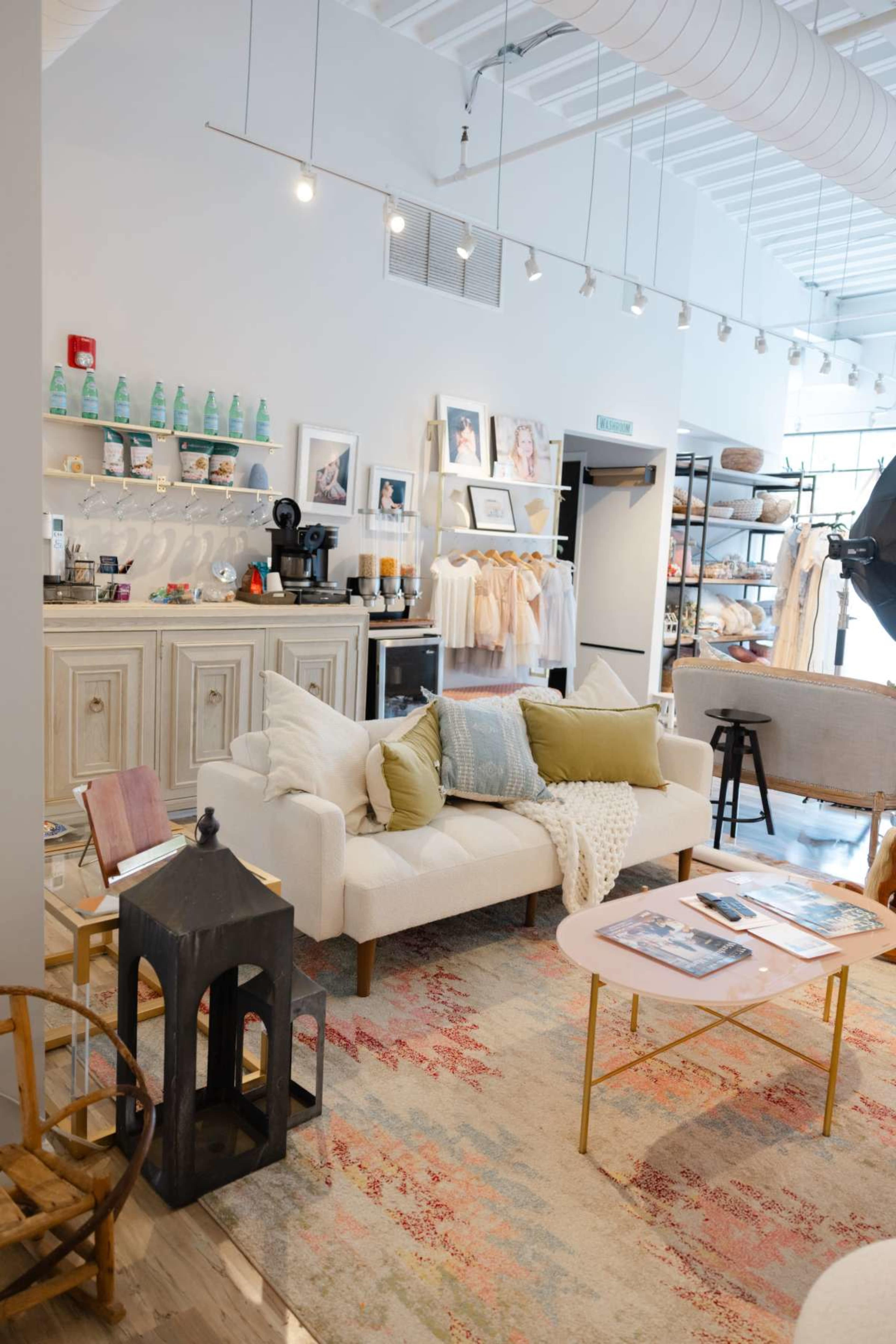 The image shows a cozy boutique interior featuring a white sofa with decorative pillows, a rug, and shelves displaying various items.
