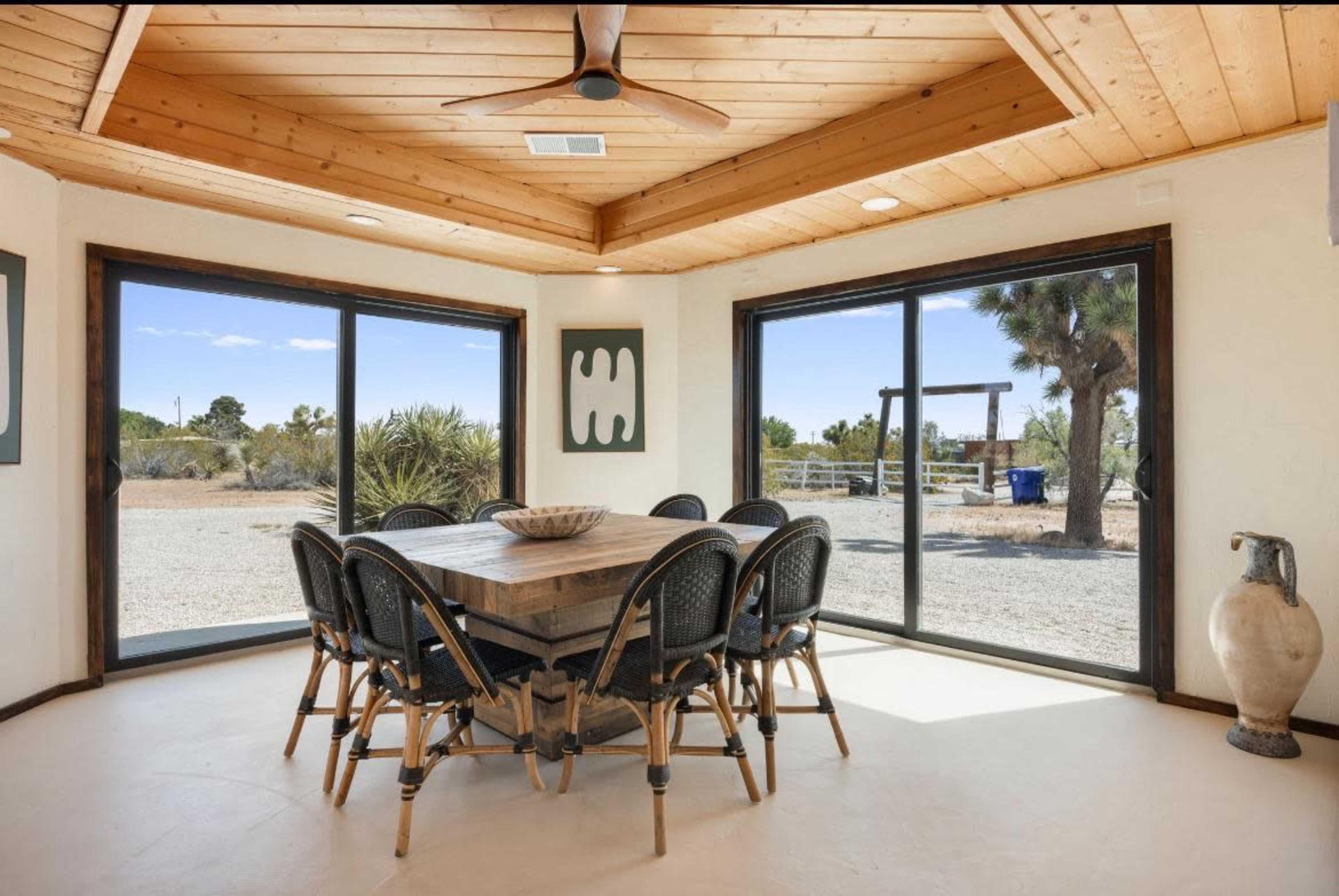 A spacious dining area features a wooden table surrounded by wicker chairs, with large windows offering a view of the outdoors.
