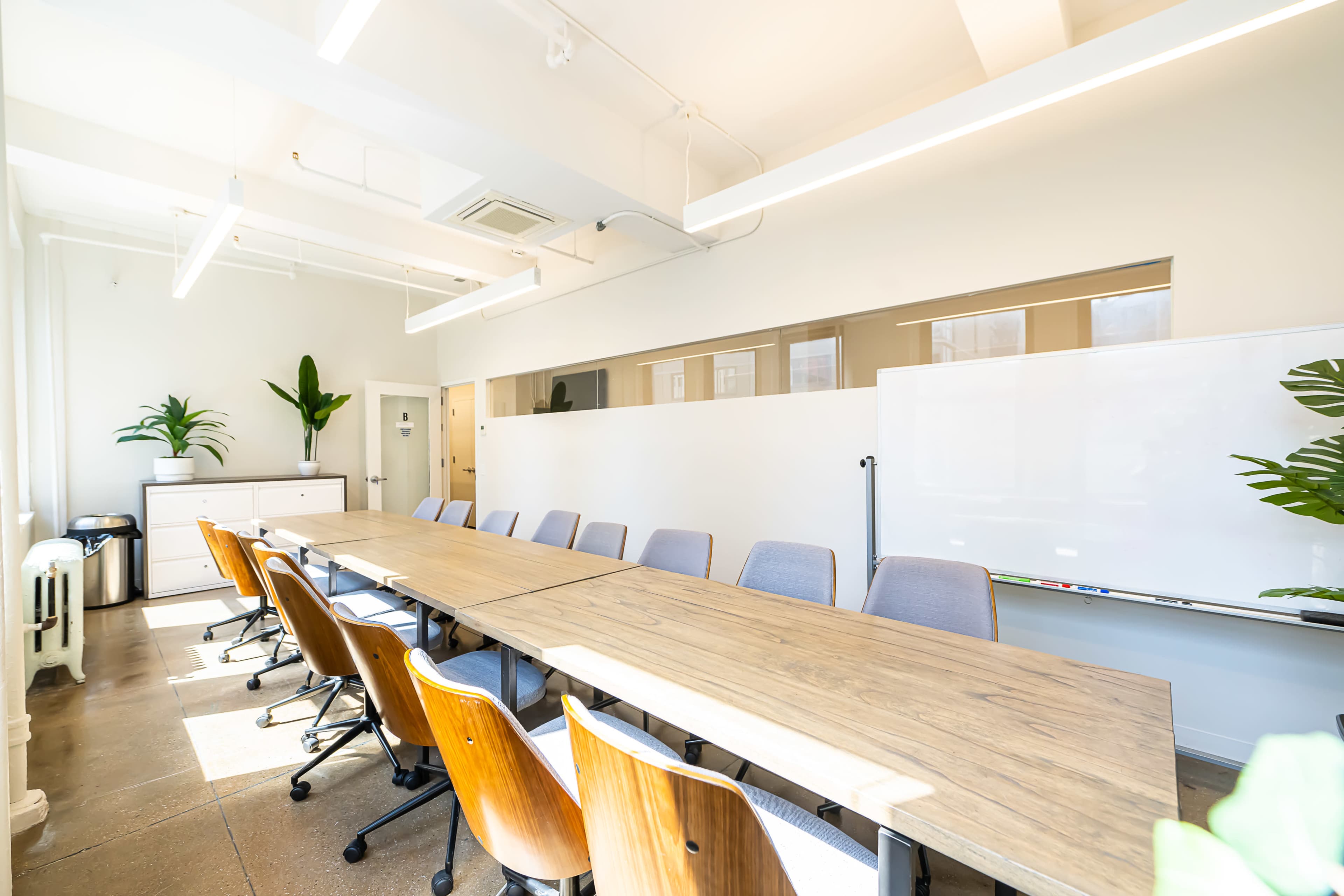 A bright conference room with a long wooden table surrounded by chairs, plants in the corners, and a whiteboard on the wall.