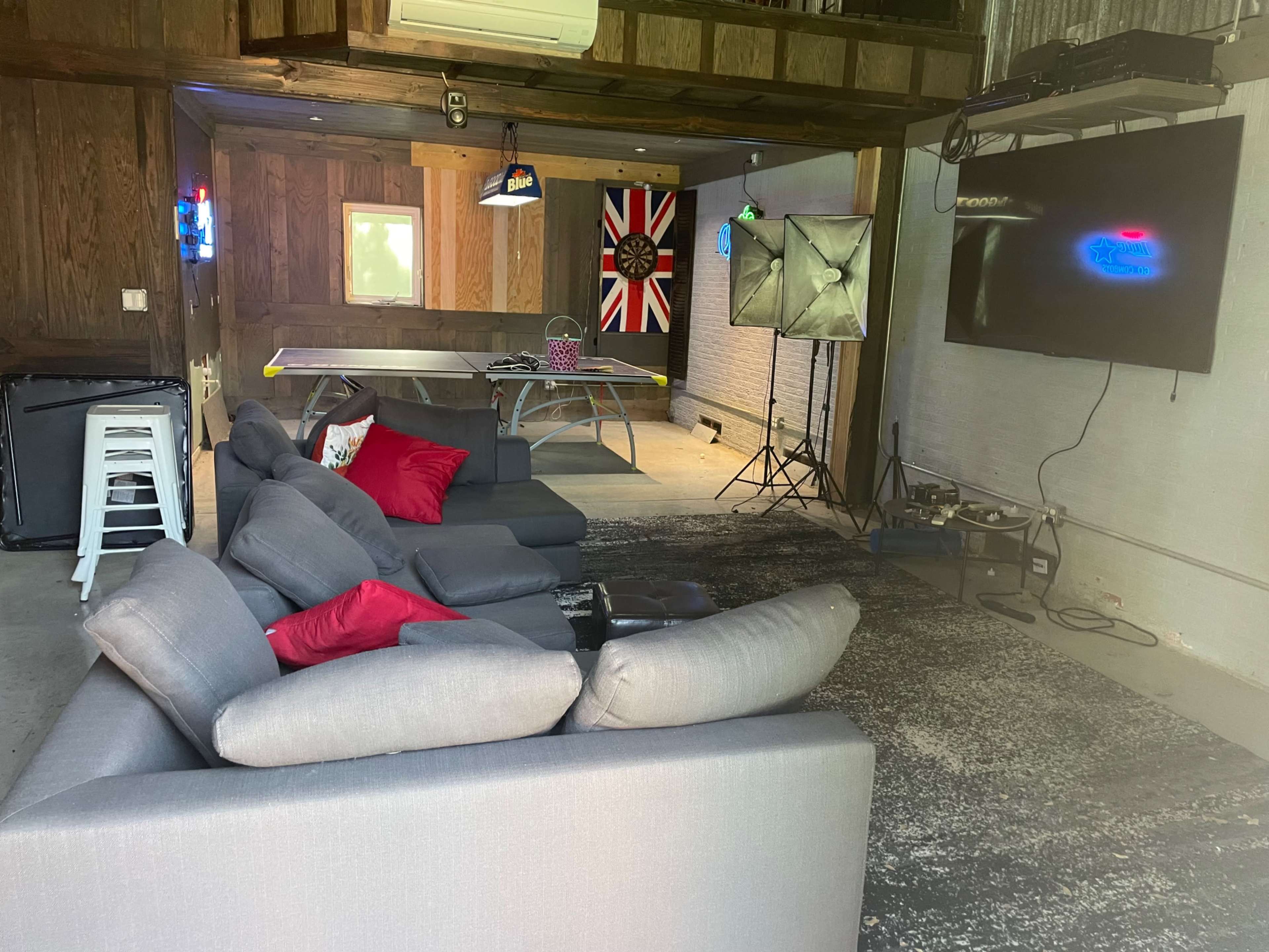 The image shows a cozy, dimly lit recreational room featuring a comfortable seating area with gray sofas, a ping pong table, and a large TV mounted on the wall.