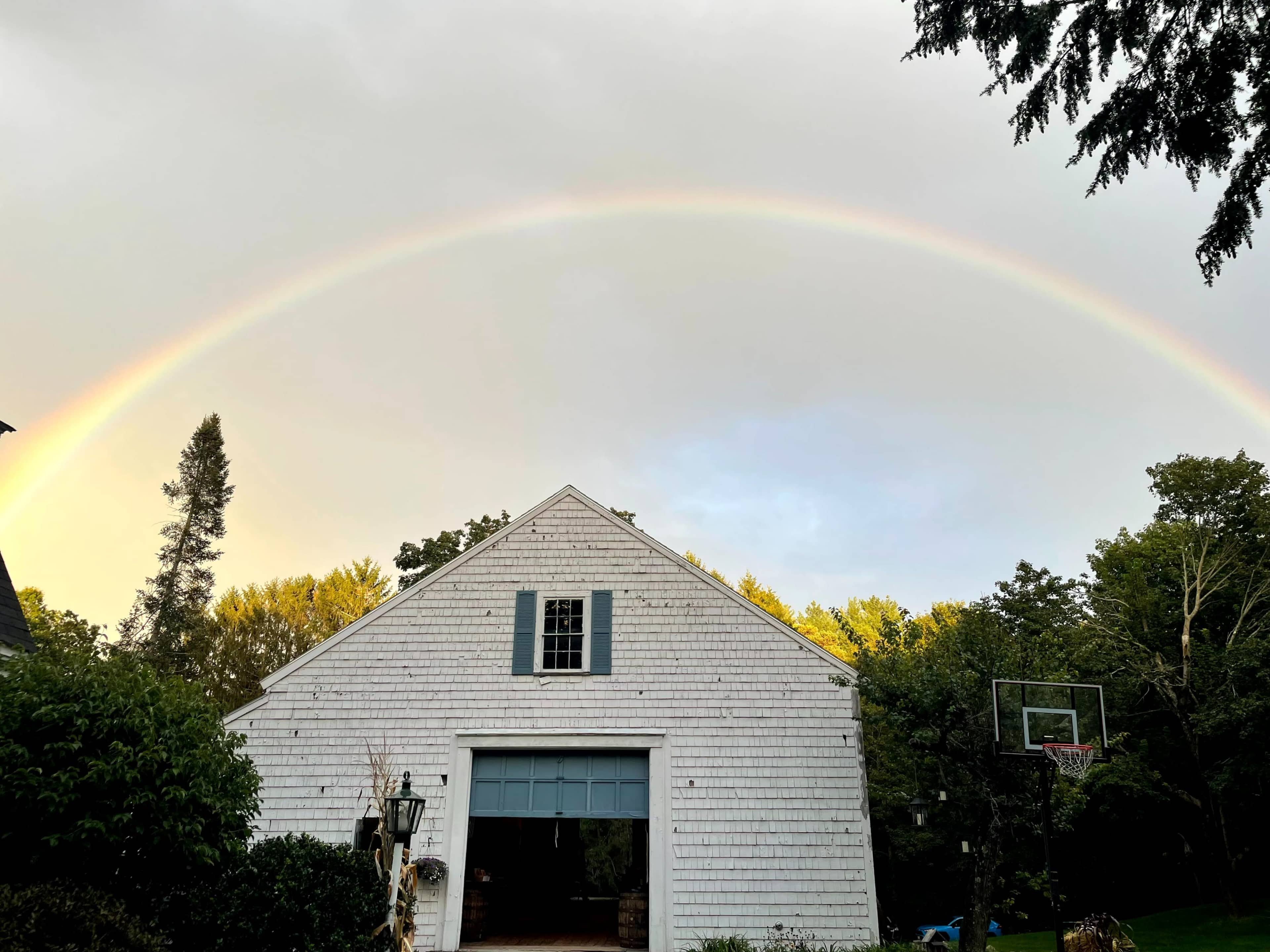 A rainbow arcs over a white barn-style garage surrounded by trees and a basketball hoop.