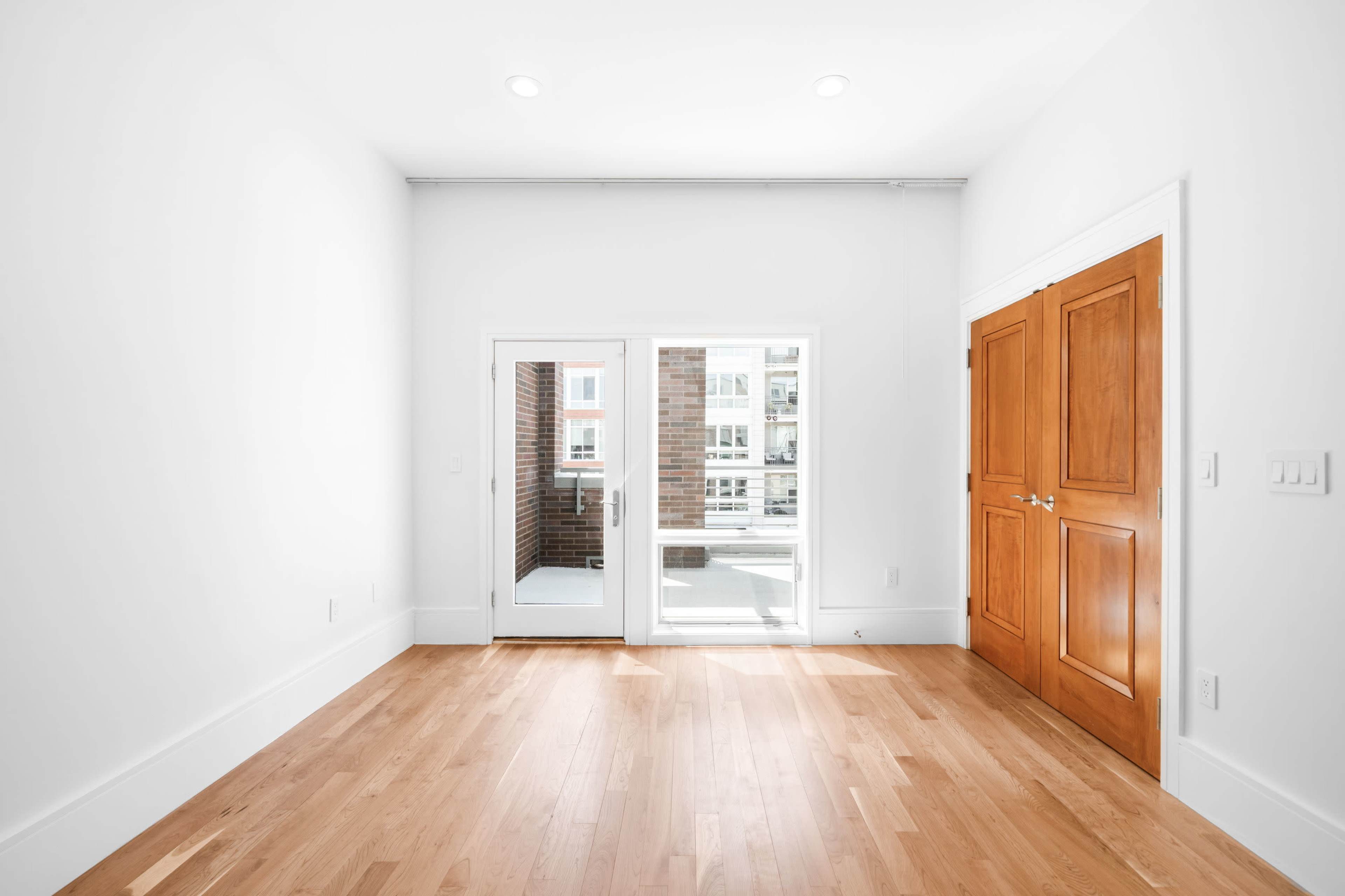 The image shows a bright, empty room with light wood flooring and large glass doors leading to a balcony.