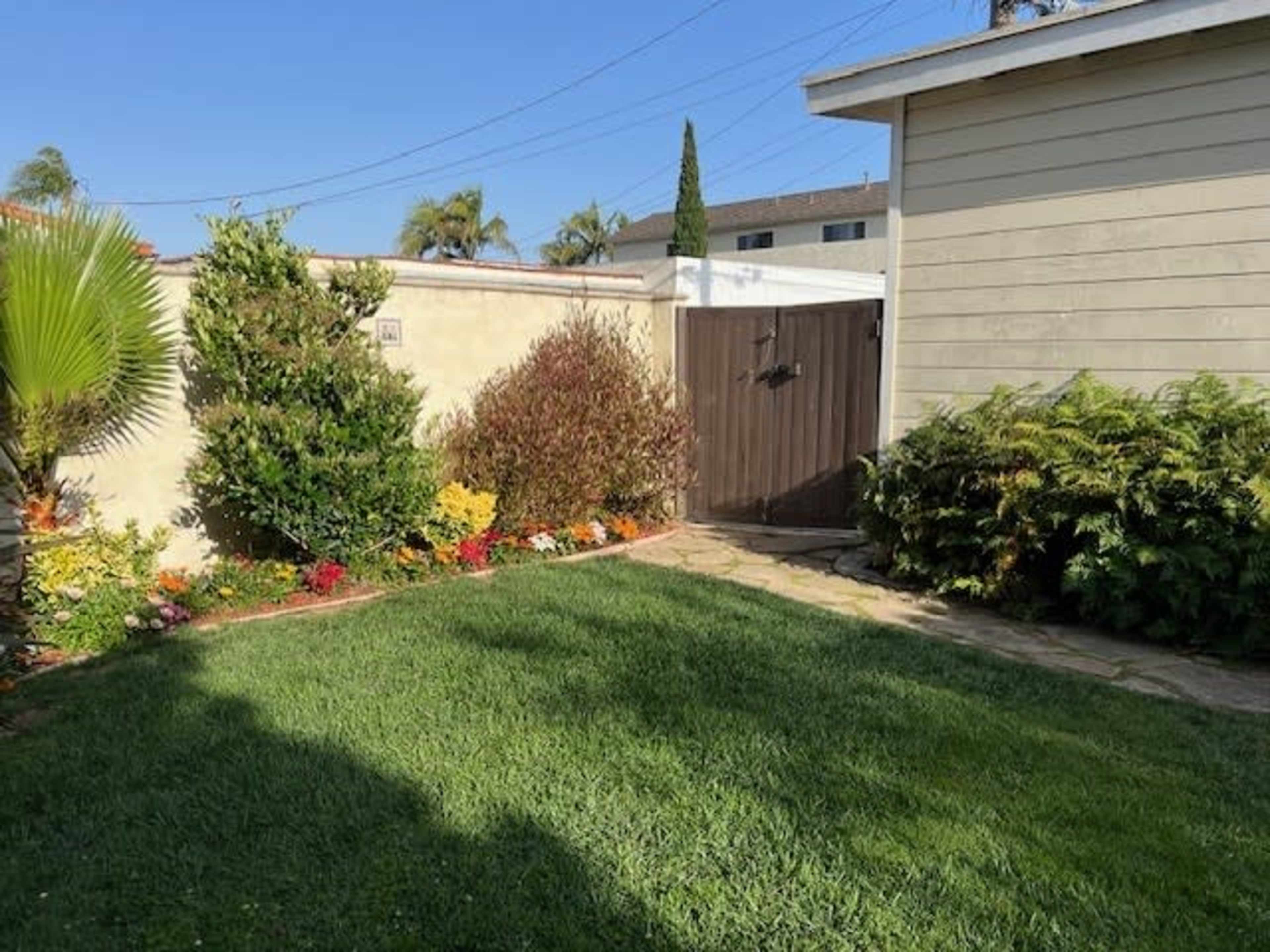 A well-maintained garden features a grassy area surrounded by colorful flowers and shrubs, adjacent to a wooden gate and a house.