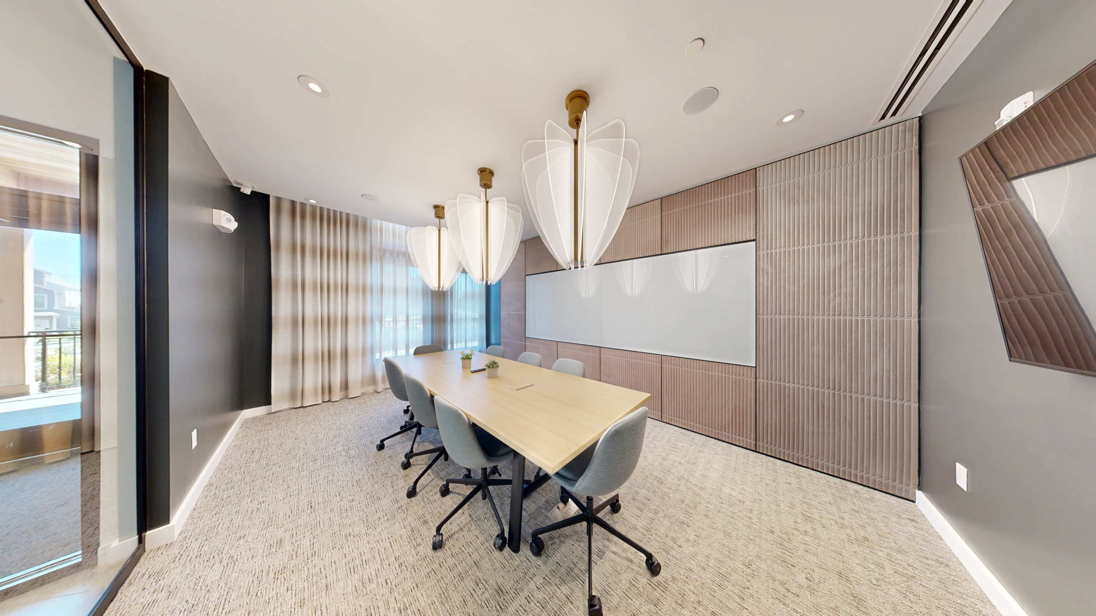 A modern conference room features a long wooden table surrounded by rolling chairs, with large pendant lights hanging from the ceiling and a whiteboard on the wall.