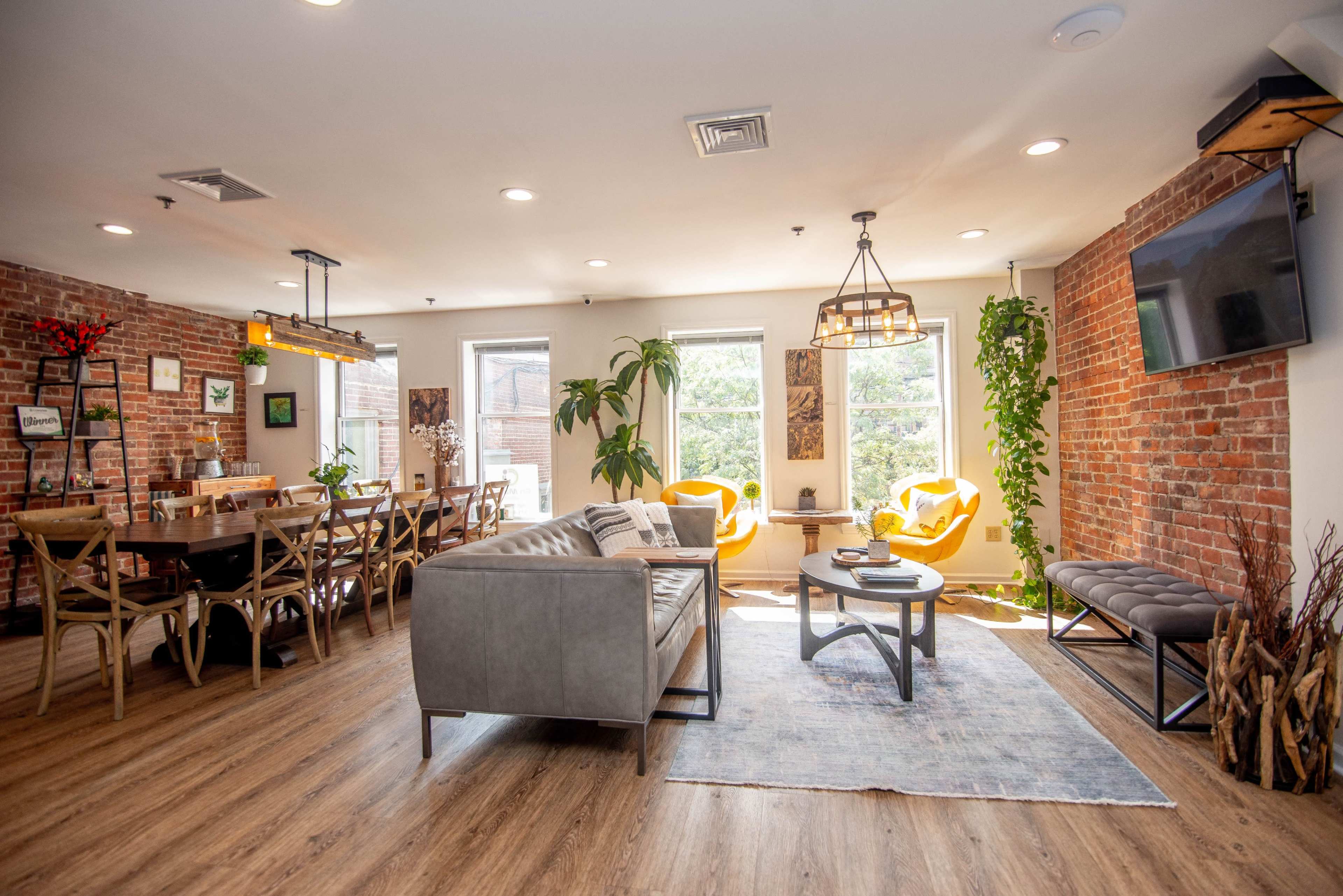 A modern lounge area with a gray sofa, a wooden dining table, bright yellow armchairs, and brick walls, illuminated by natural light from the windows.