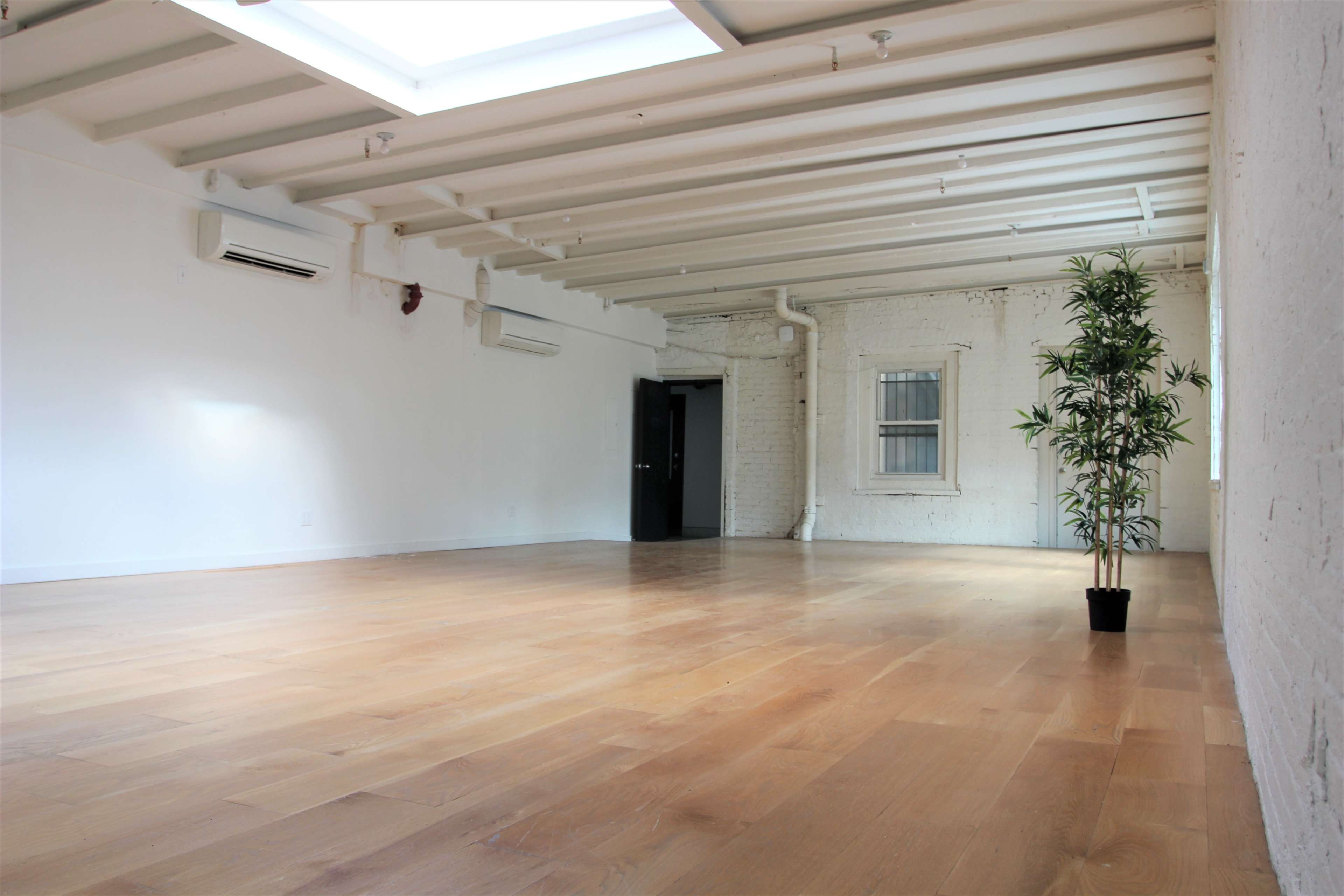 A spacious, empty room with wooden flooring, exposed brick walls, and a small potted plant in one corner.