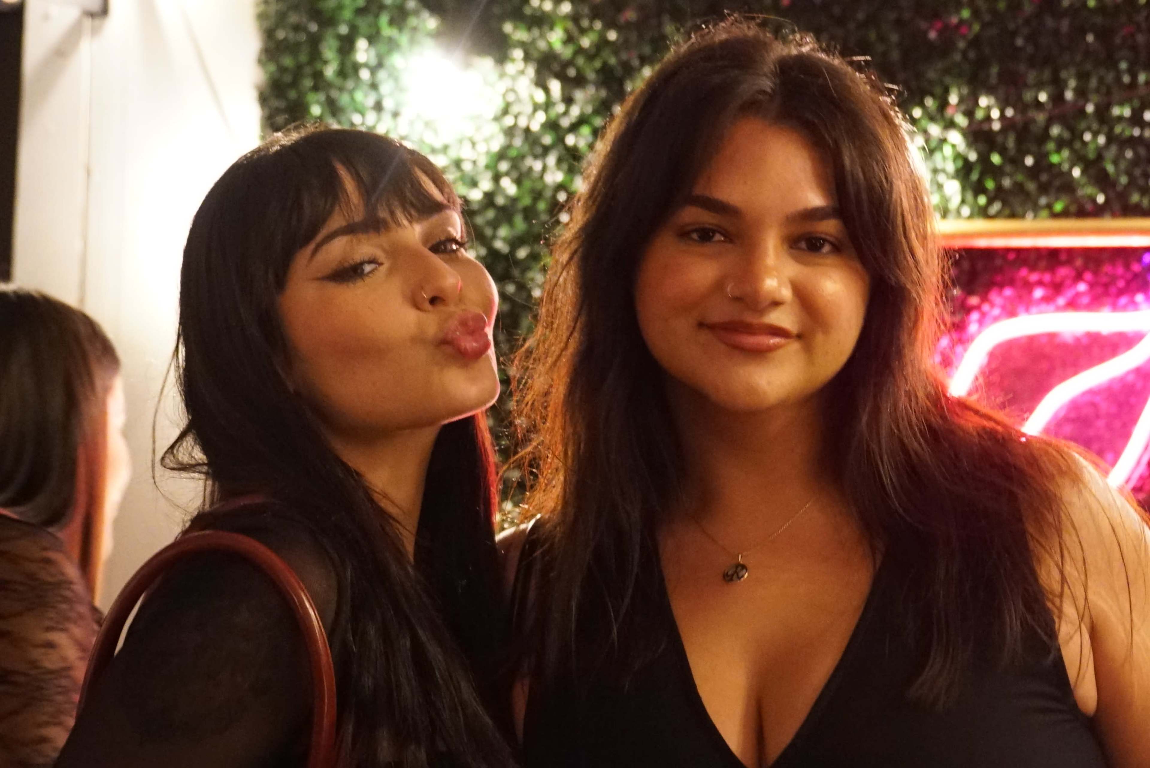 Two women pose for a photo against a backdrop of greenery and neon lights.
