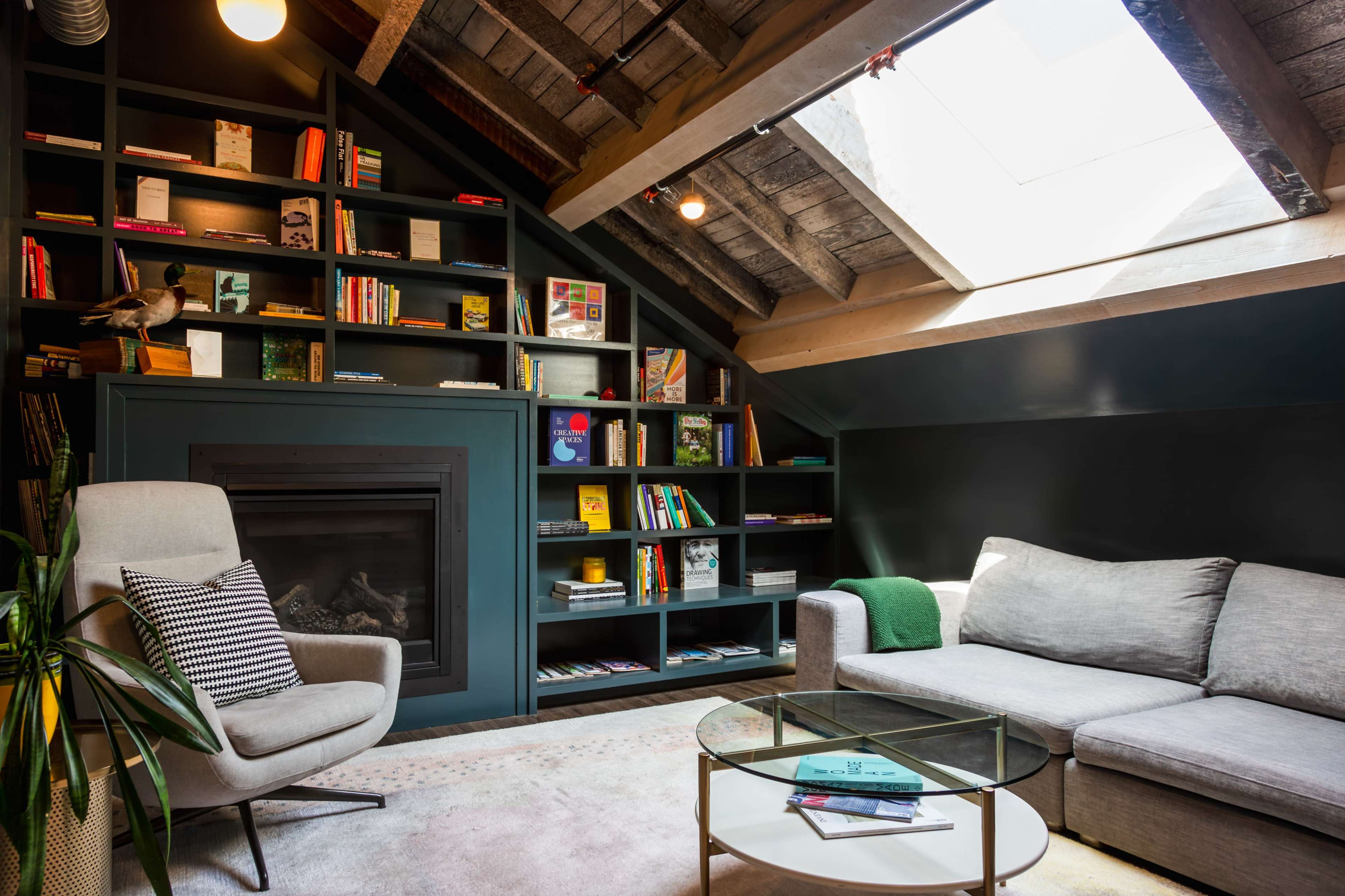 A cozy reading nook features a gray sofa, a modern armchair, and a bookshelf filled with books, all under a skylight in a rustic attic space.