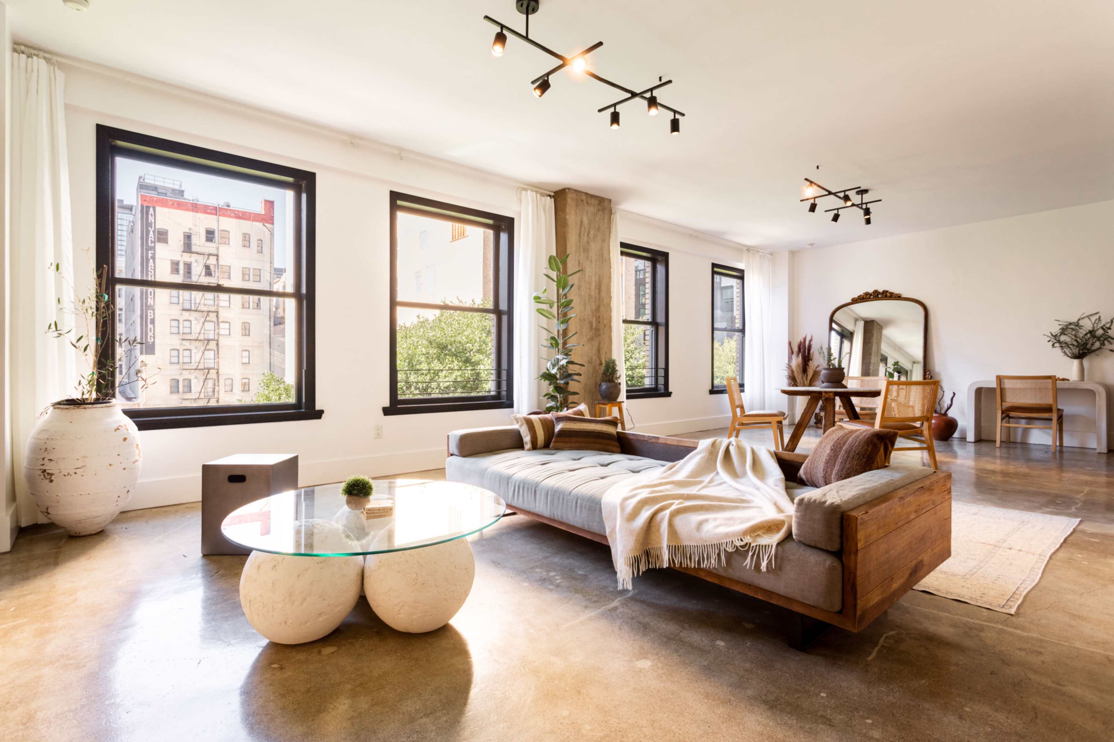 A spacious, modern living area with large windows, a wooden sofa, a glass coffee table, and a dining area featuring a mirror and wooden chairs.