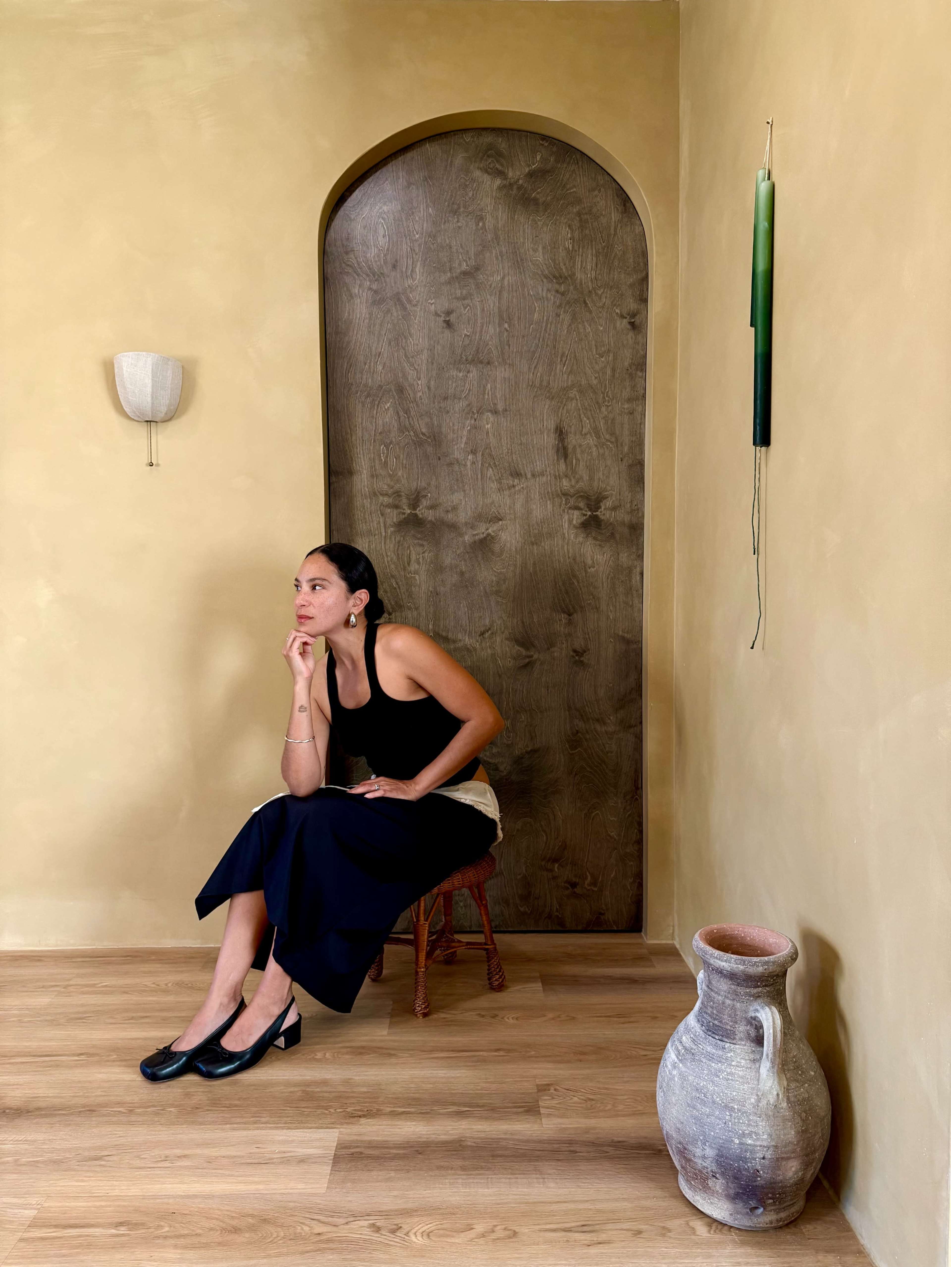 A woman sits on a wooden stool beside a rustic pottery jar, positioned in a warmly lit interior with a curved door and simple decor.