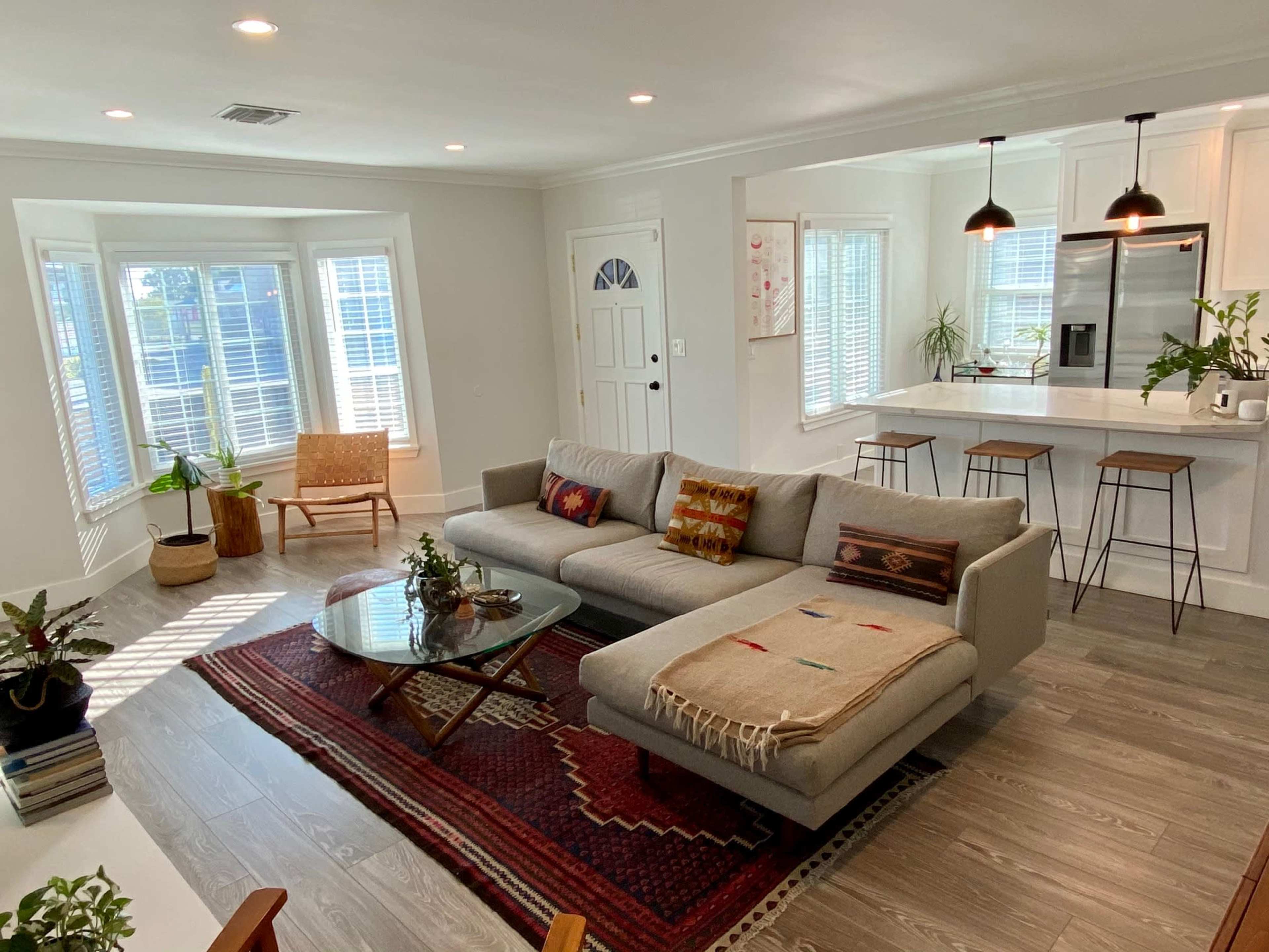 A modern living room with a gray sectional sofa, a round glass coffee table, and a dining area featuring bar stools and a kitchen in the background.