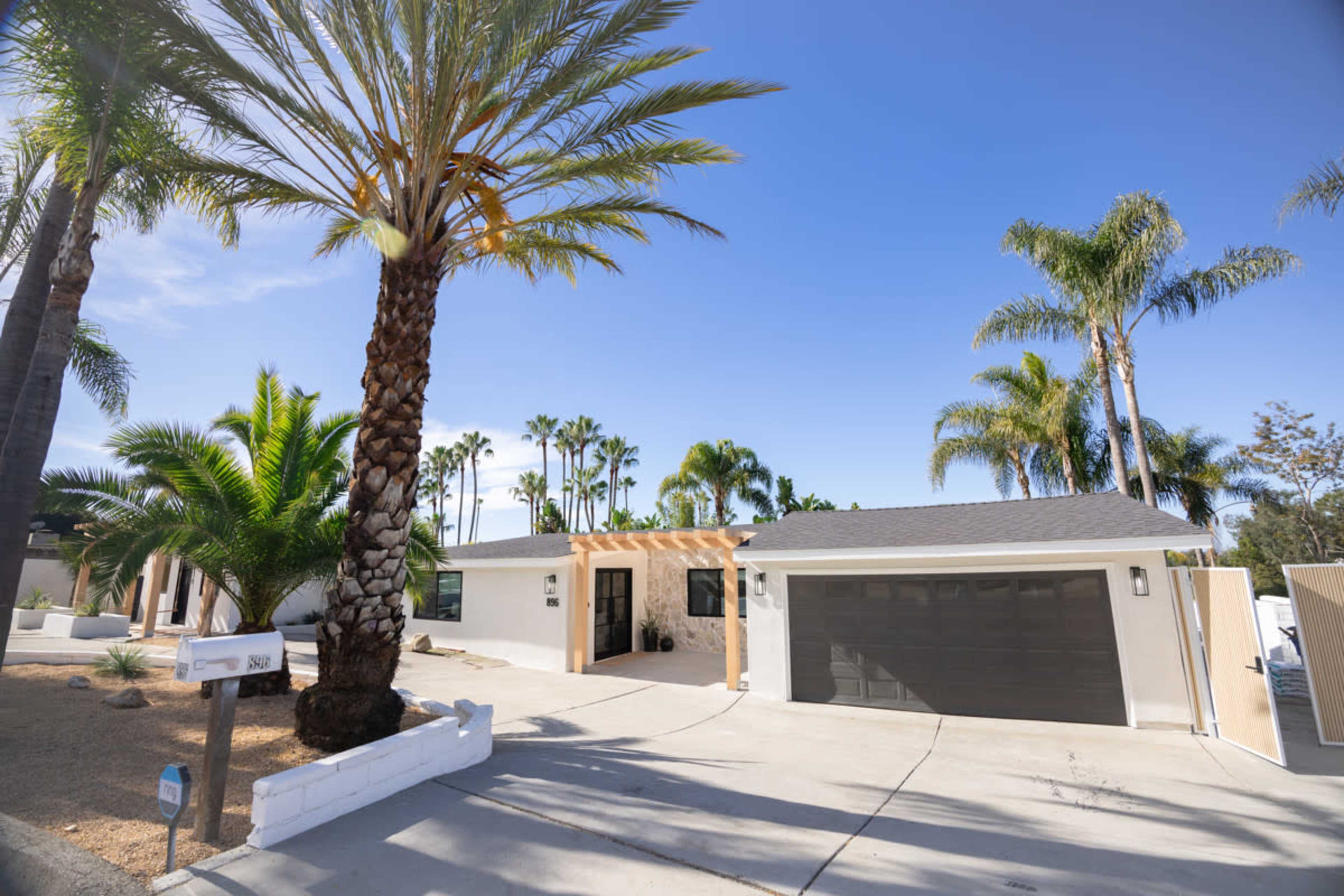 A modern single-story house with a grey roof and a two-car garage is surrounded by palm trees and landscaped vegetation.