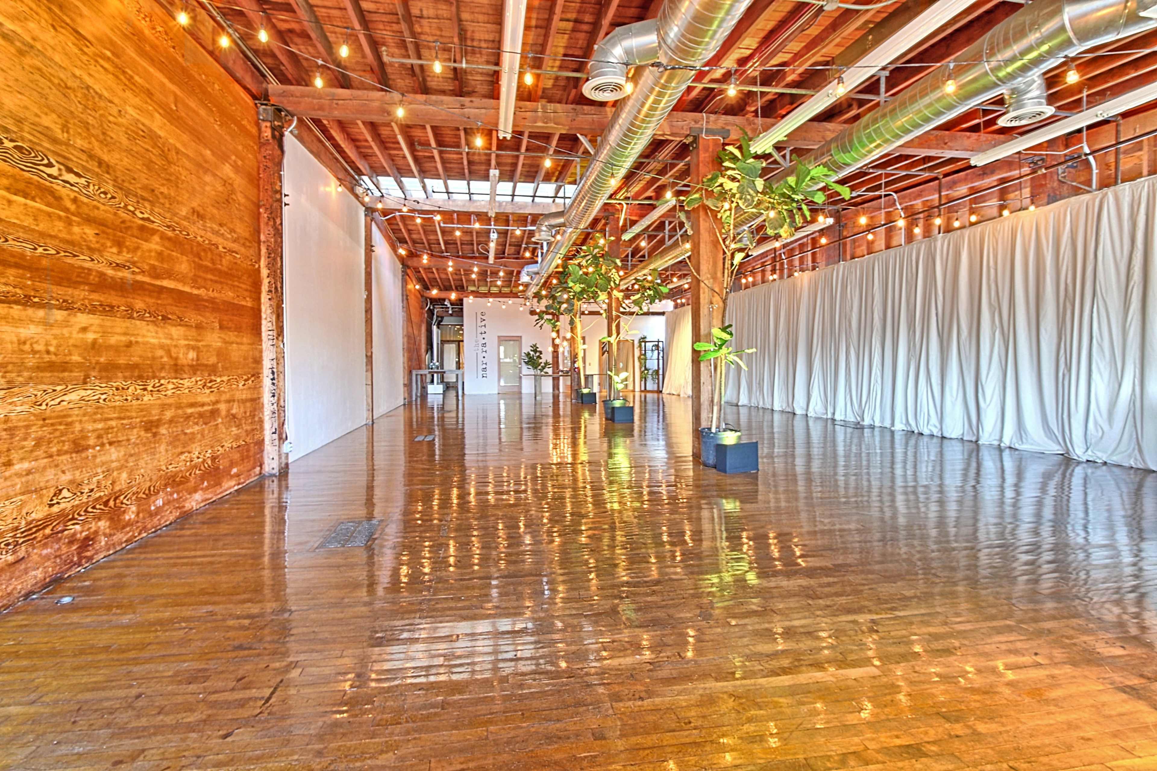 A spacious event venue with polished wooden floors, exposed wooden beams, and potted plants along the sides.