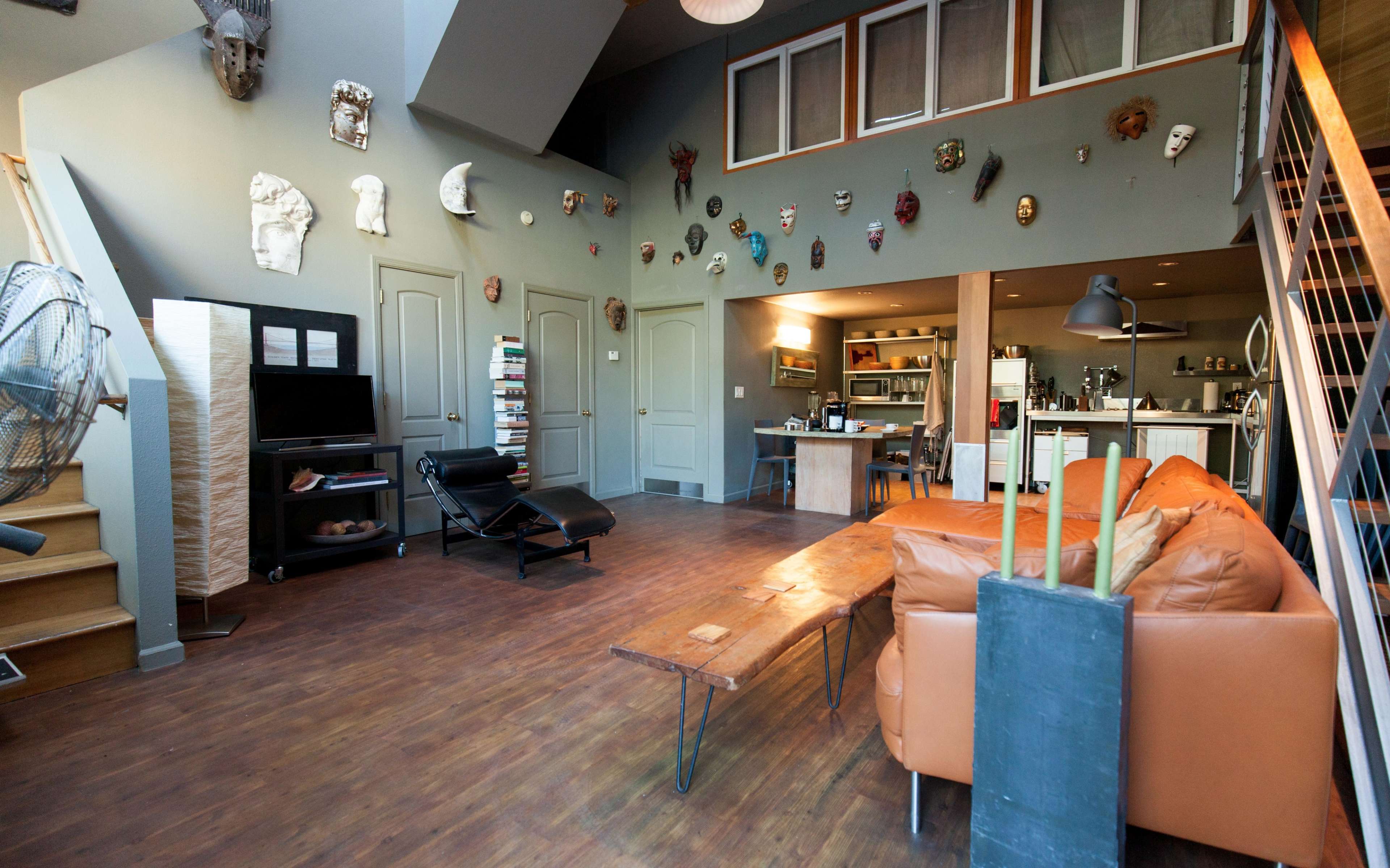 A spacious living area with wooden flooring, a brown leather couch, a coffee table, and a wall adorned with various decorative masks.