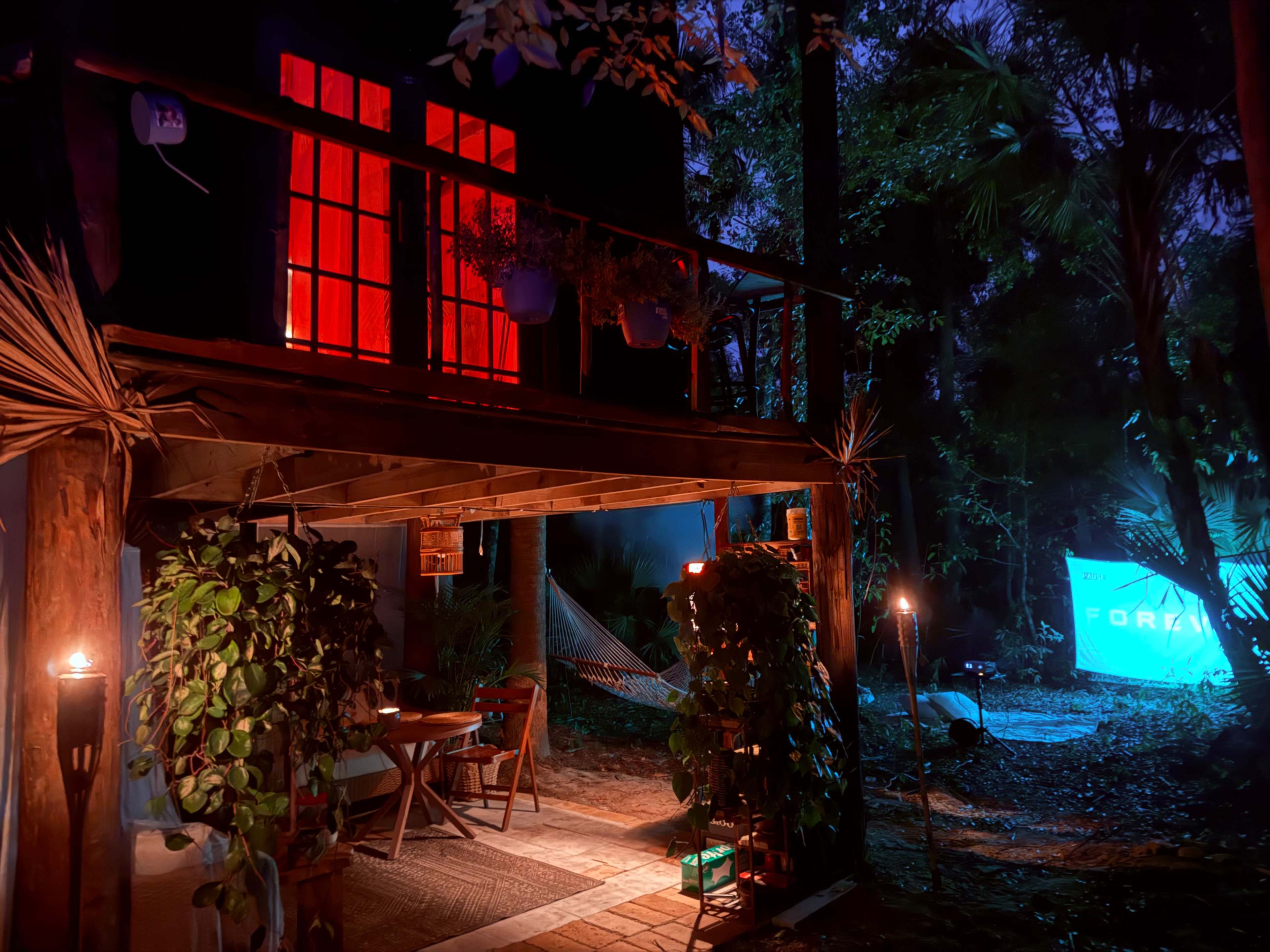 The scene depicts a cozy outdoor area with a wooden structure featuring red-lit windows, surrounded by trees and illuminated by blue light from a nearby screen.