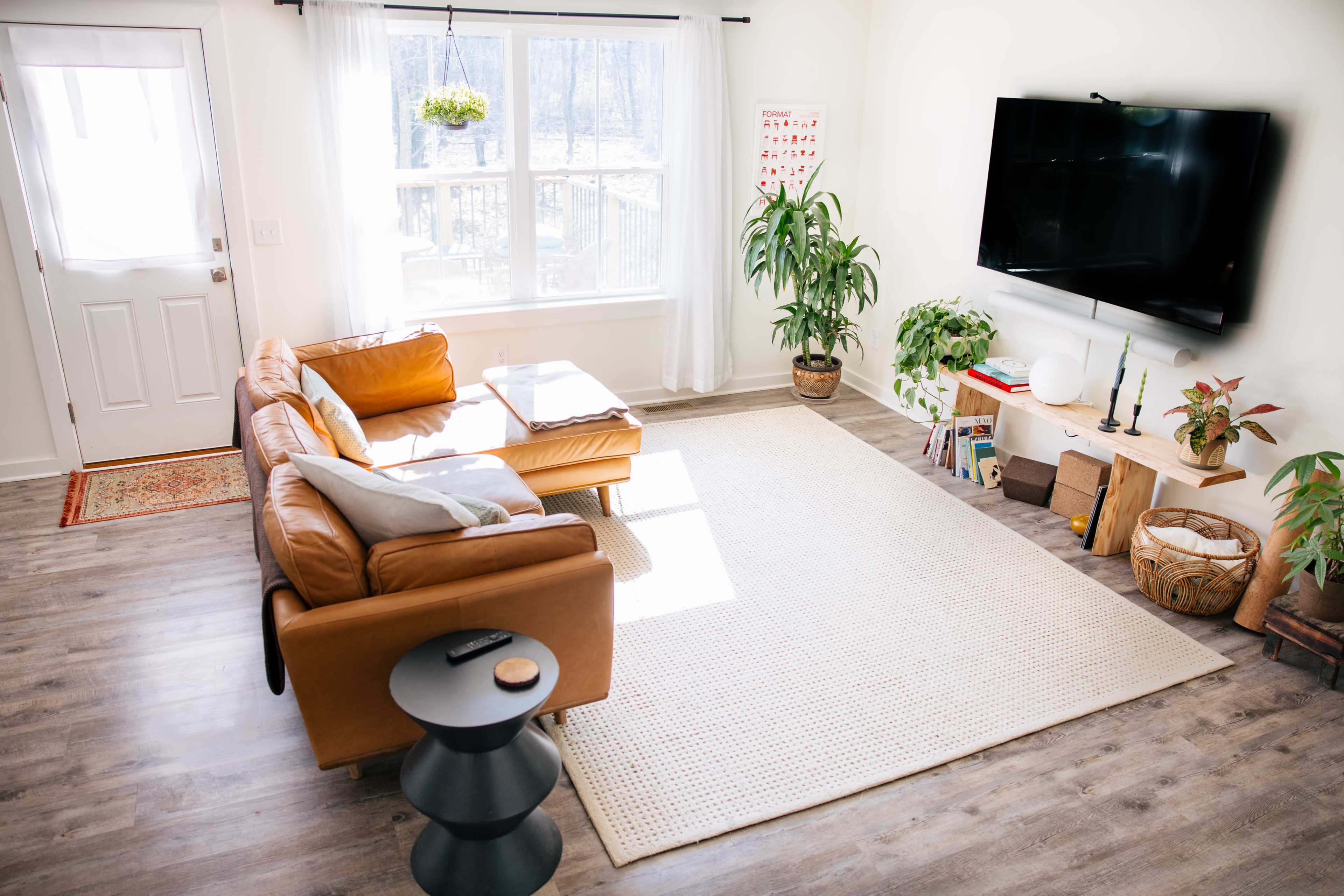 A well-lit living room features a brown leather couch, a television mounted on the wall, and various plants, with a door leading outside and a window showcasing a view of greenery.