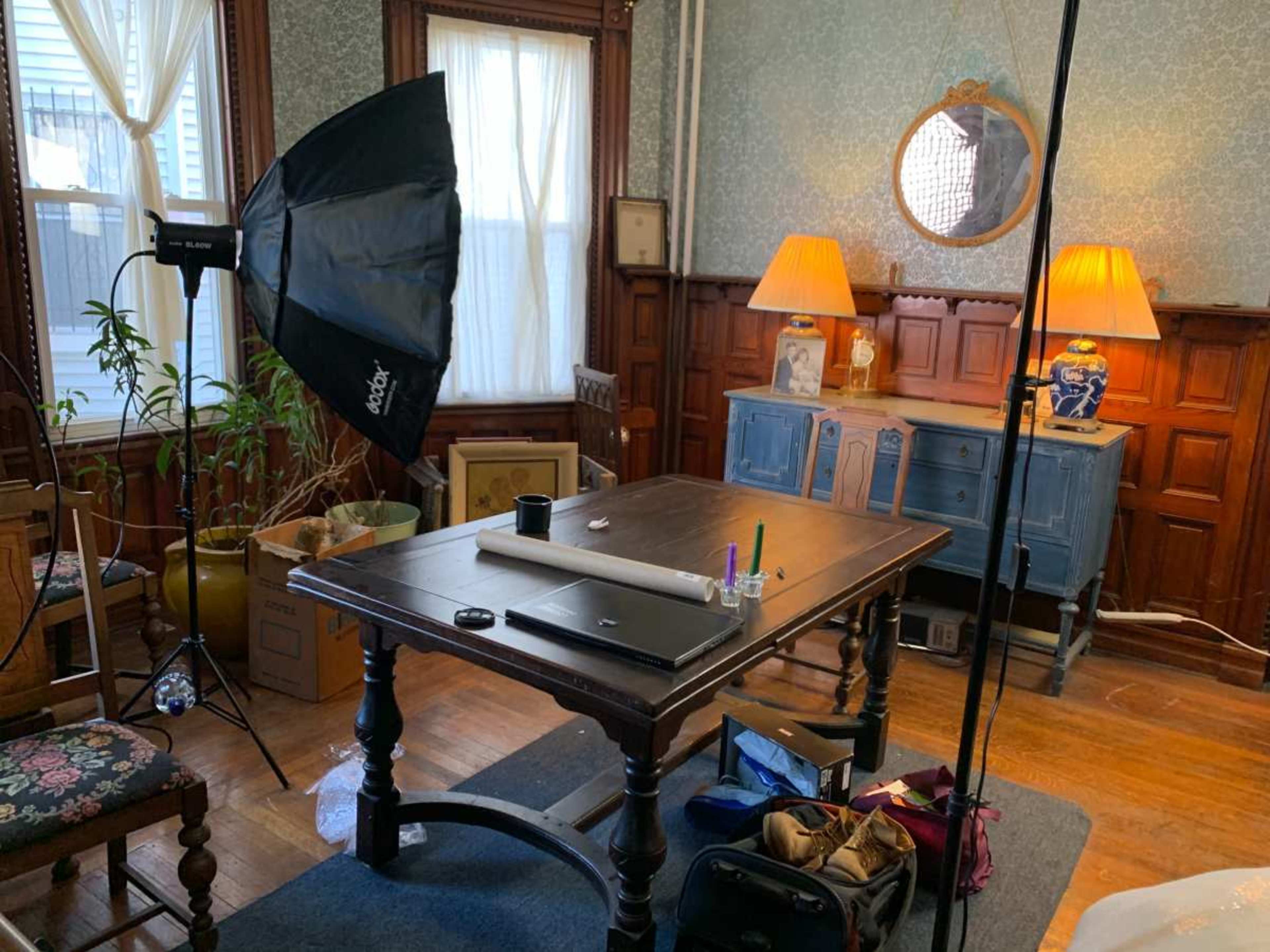 A well-furnished room features a wooden table at the center, with a photography light setup, two lamps, and various items scattered around.