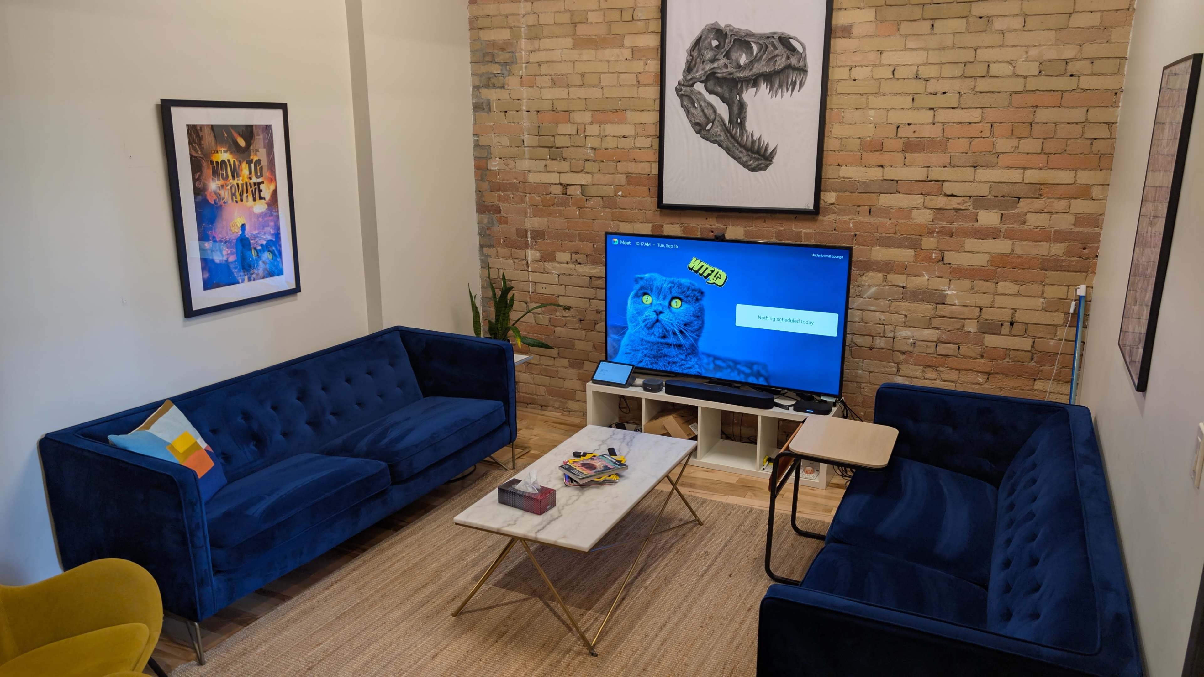 The image shows a cozy living room with two blue velvet sofas facing a coffee table, a large TV on a stand, and artwork on the exposed brick wall.