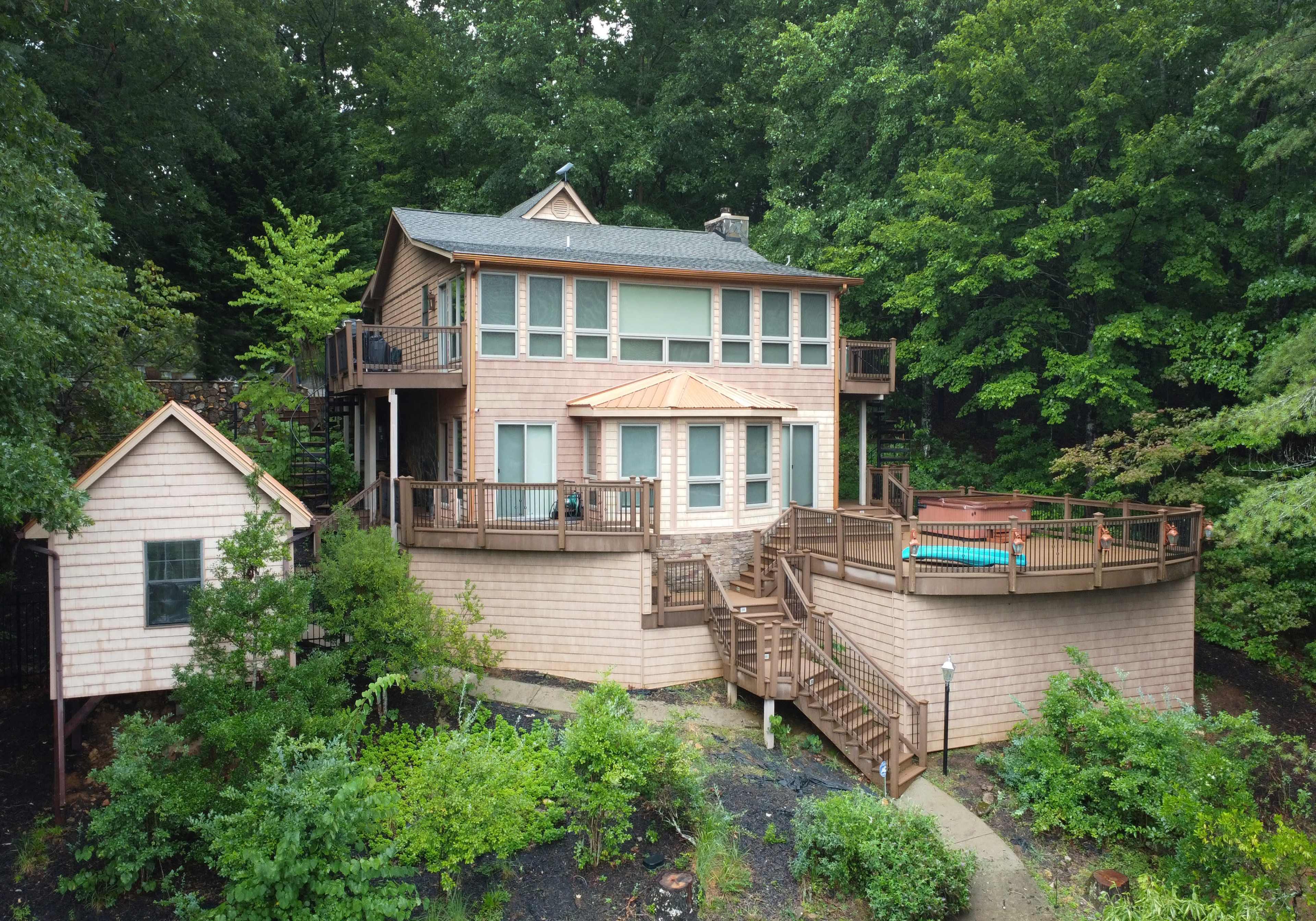 A two-story wooden house with a large deck, surrounded by greenery and trees.
