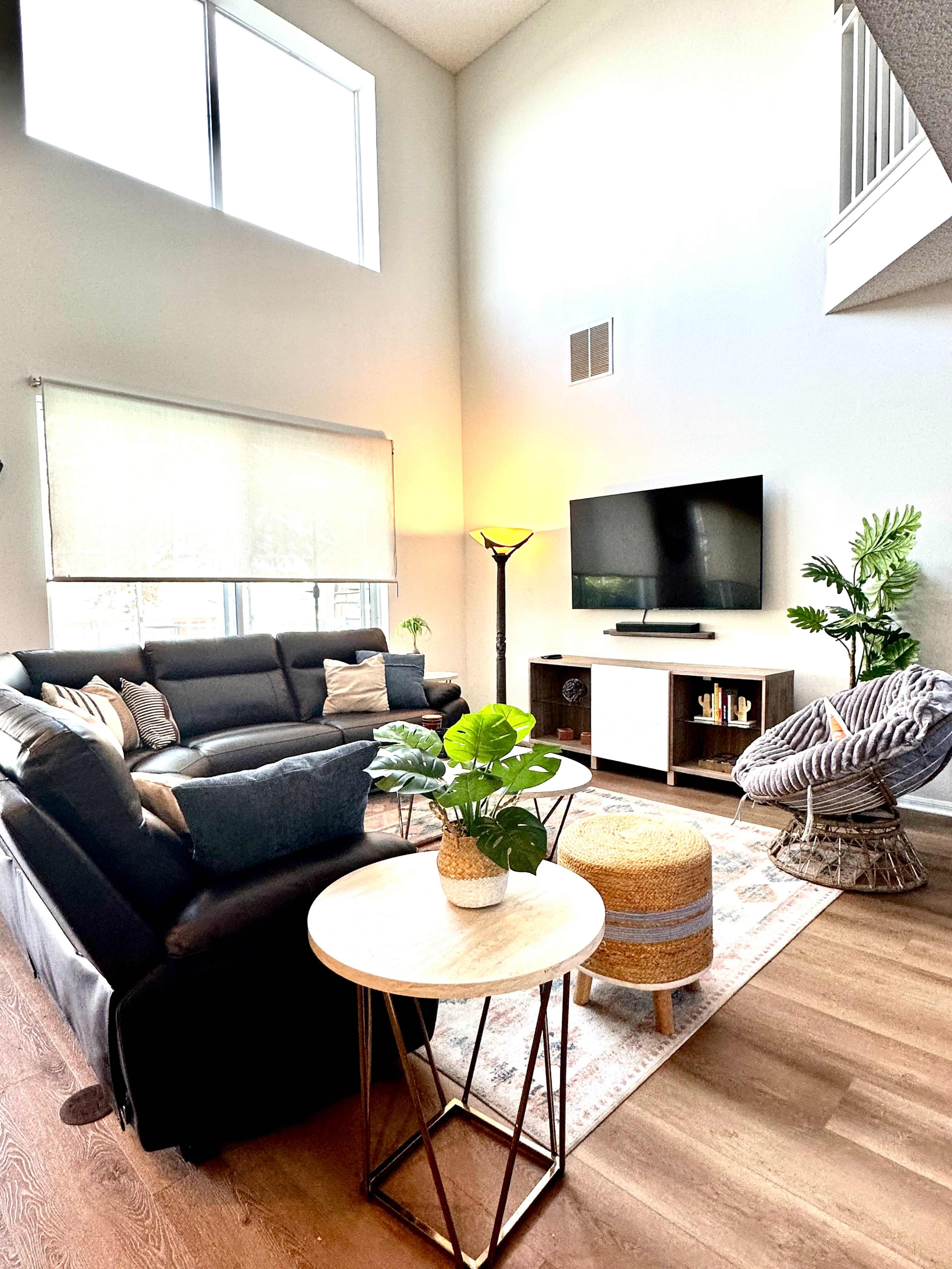 The image shows a modern living room with a black sectional sofa, a coffee table, a TV stand, and decorative plants, illuminated by natural light from tall windows.
