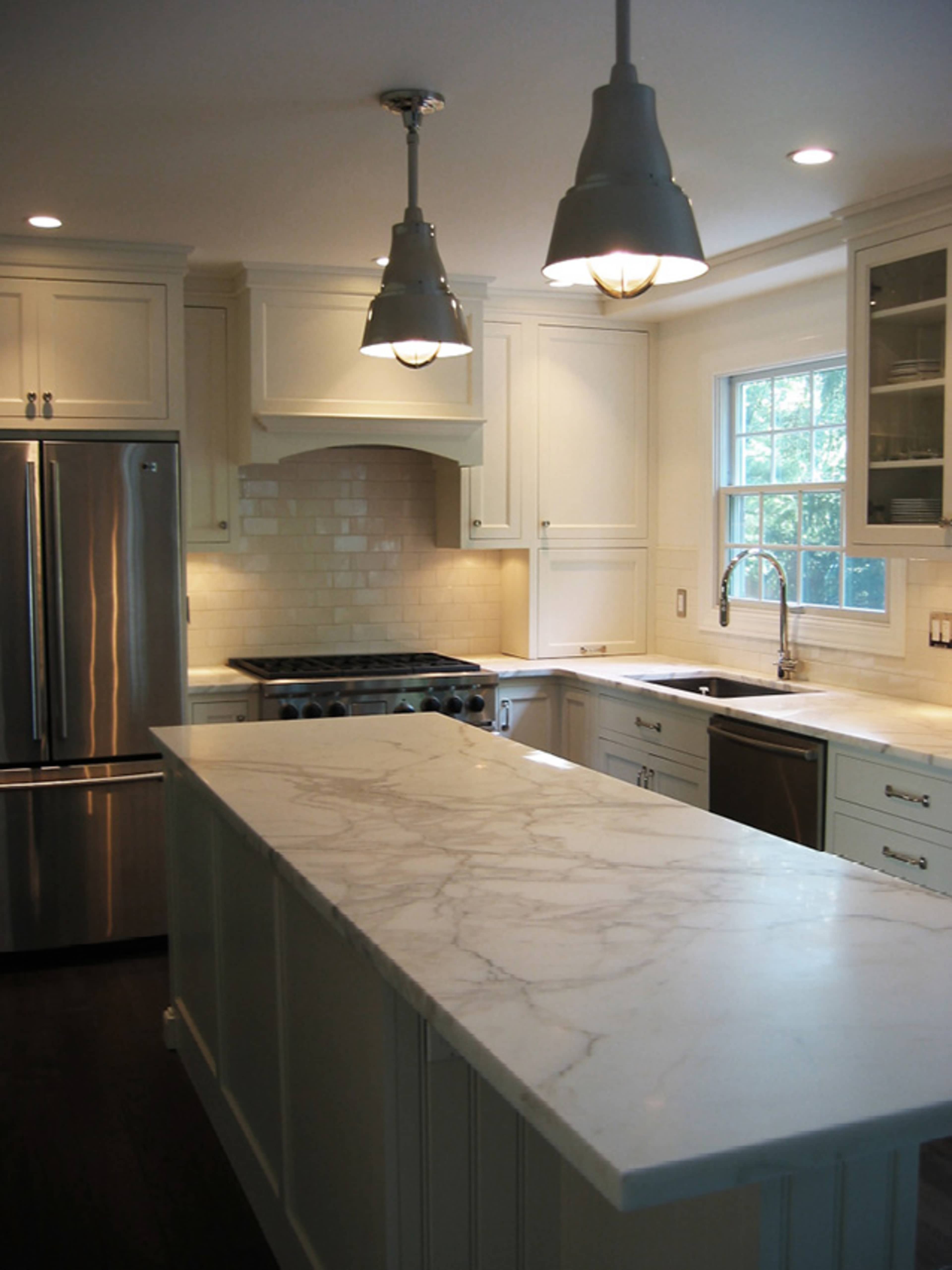 A modern kitchen featuring white cabinetry, a large marble island, stainless steel appliances, and pendant lighting.