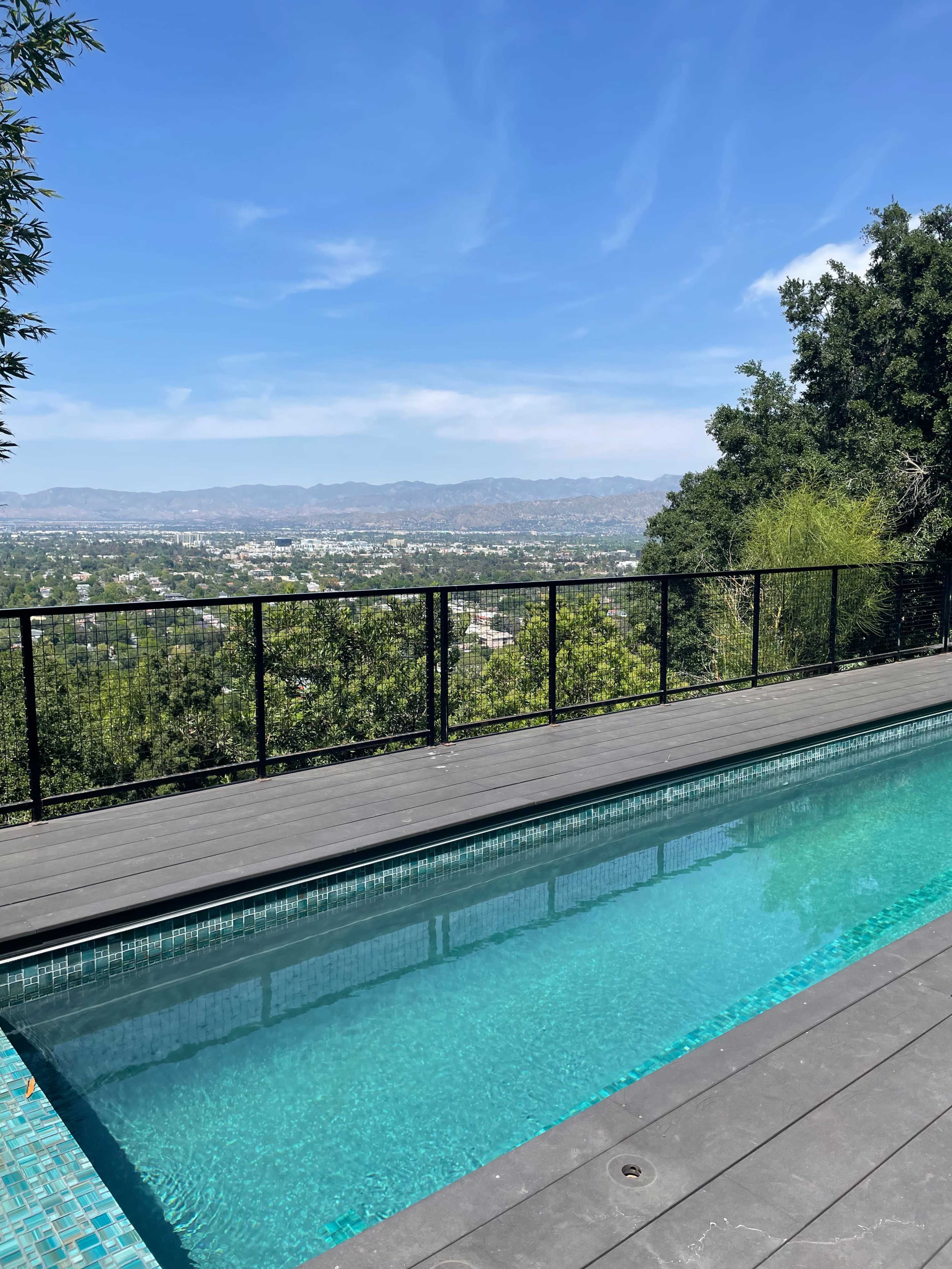 A swimming pool with a clear view of a city landscape and distant mountains is surrounded by a metal railing and greenery.