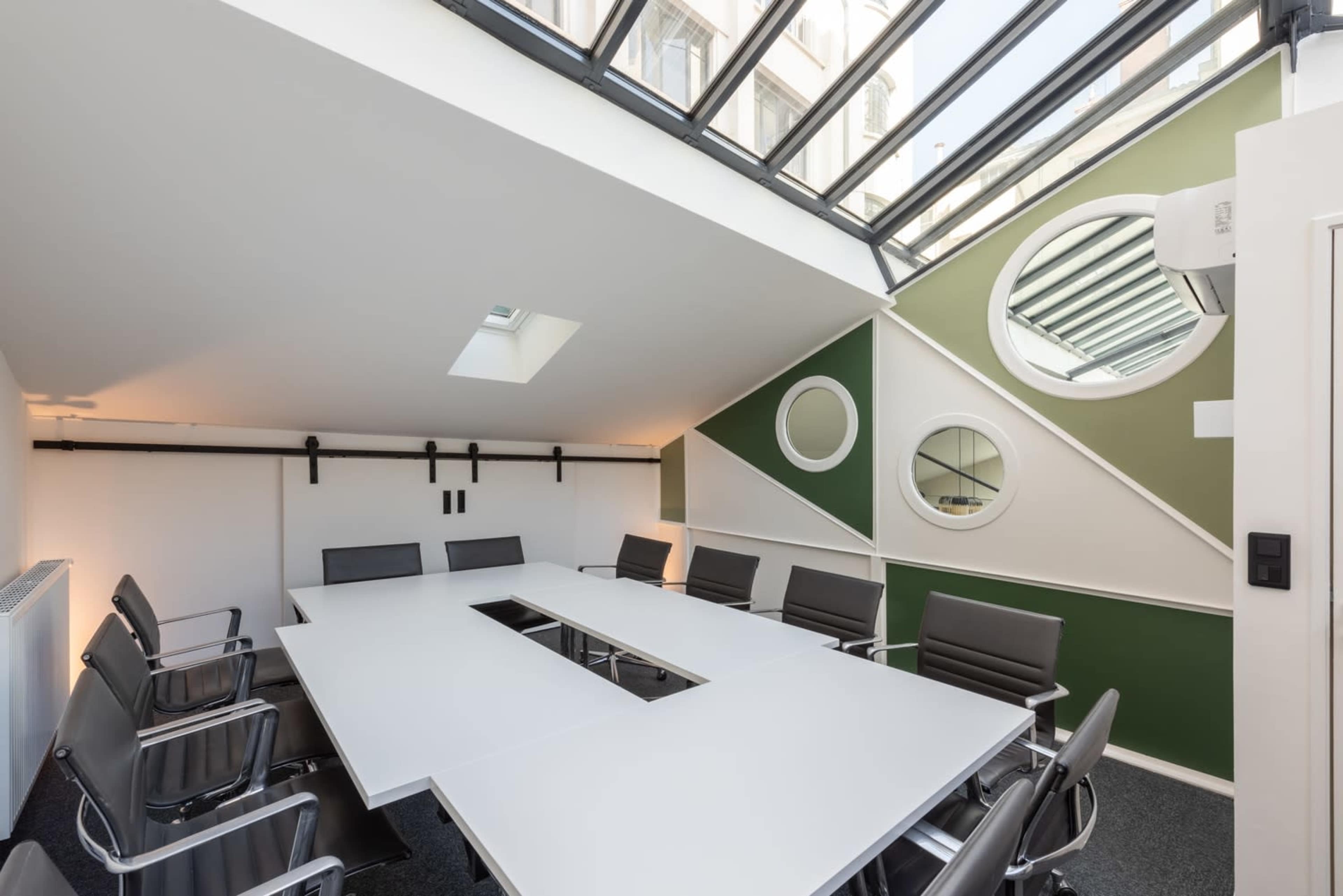 The image shows a conference room with a large white table surrounded by black chairs, set under a glass ceiling and featuring geometric green walls.