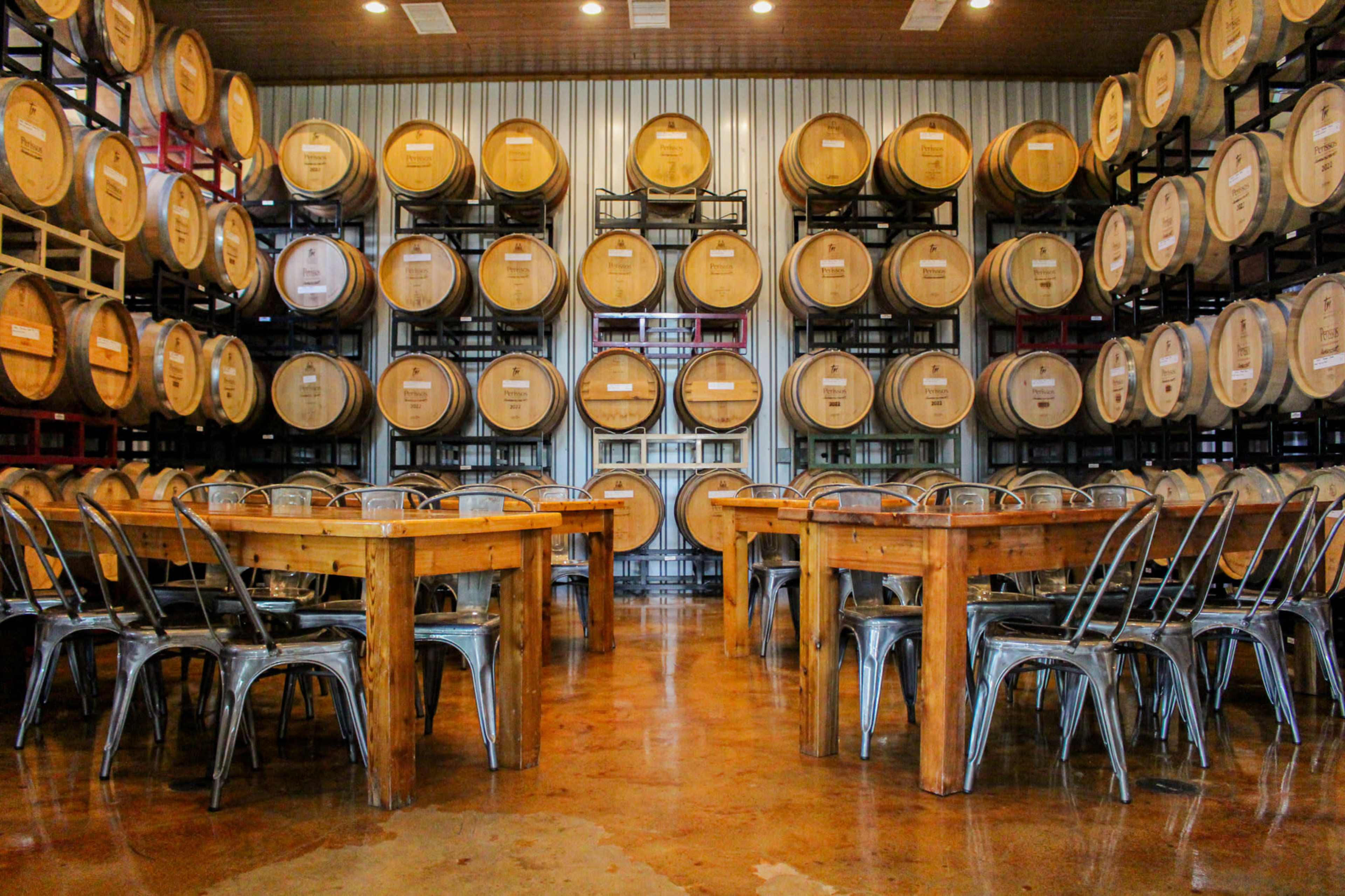A spacious tasting room features wooden tables surrounded by metal chairs, with rows of wine barrels stacked against the wall.