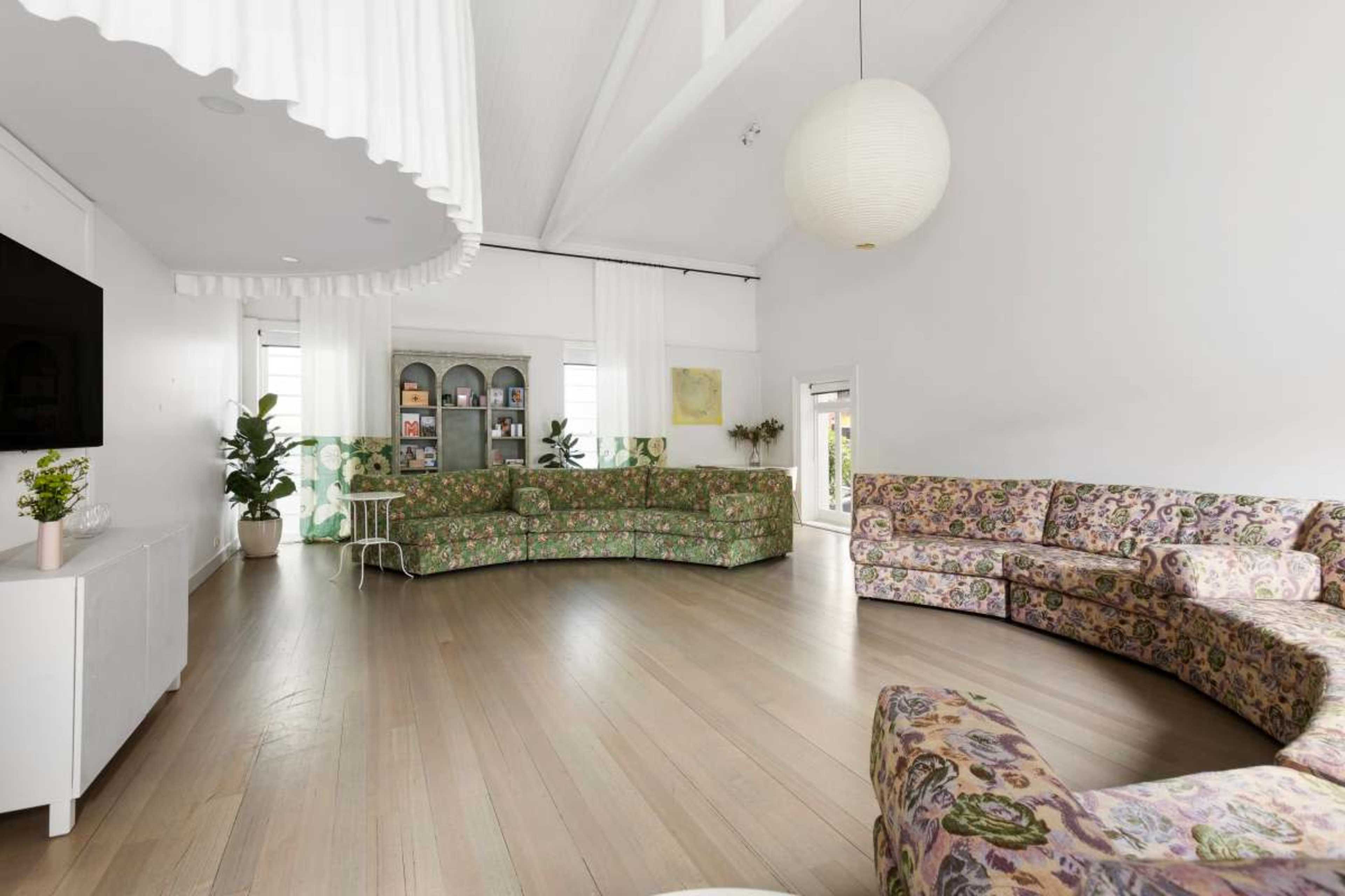The image shows a spacious room with a circular arrangement of floral-patterned sofas, large windows, and wooden flooring.