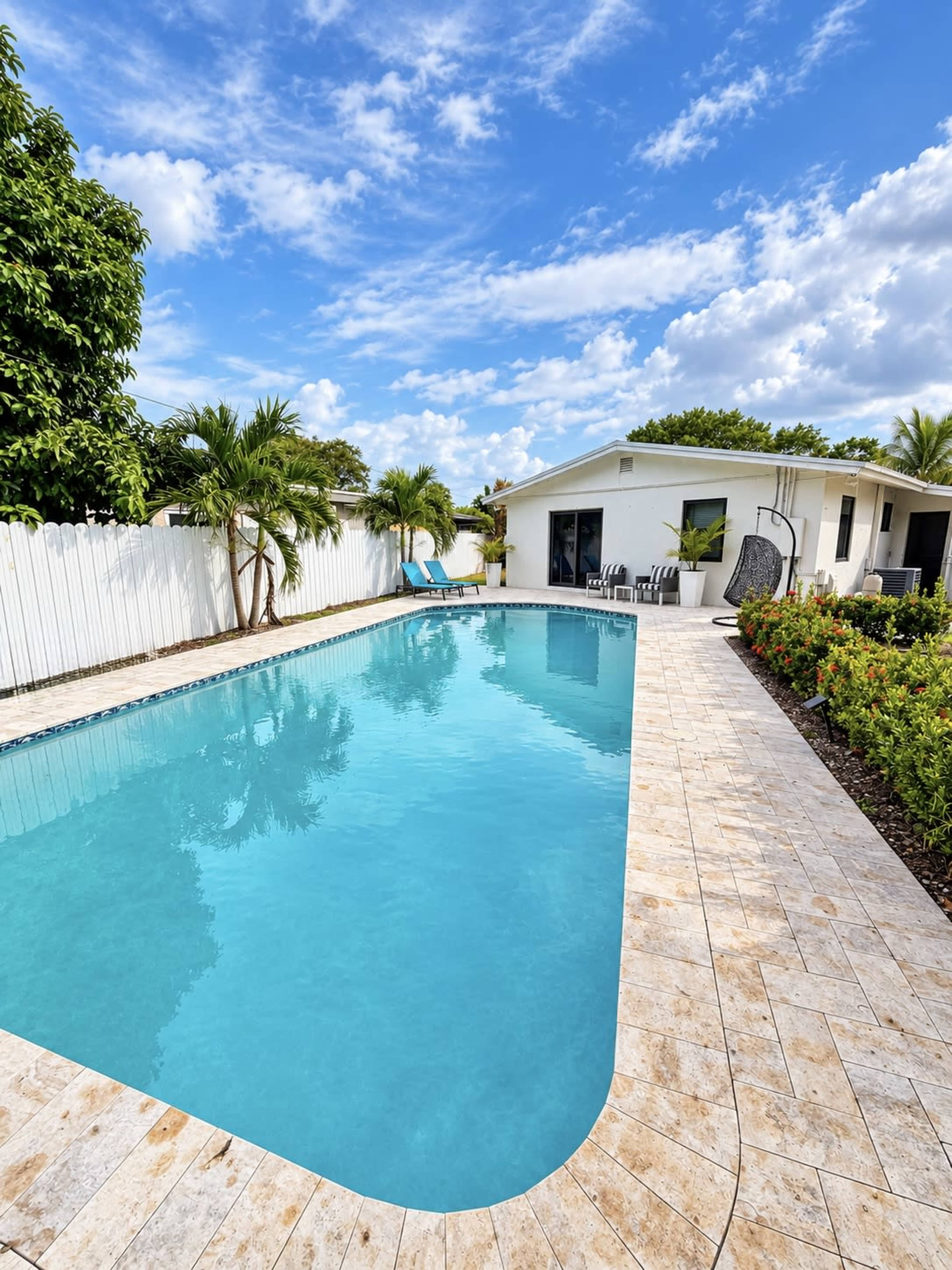 The image shows a clear, blue swimming pool surrounded by a tiled deck, tropical plants, and a white fence, with a house in the background.