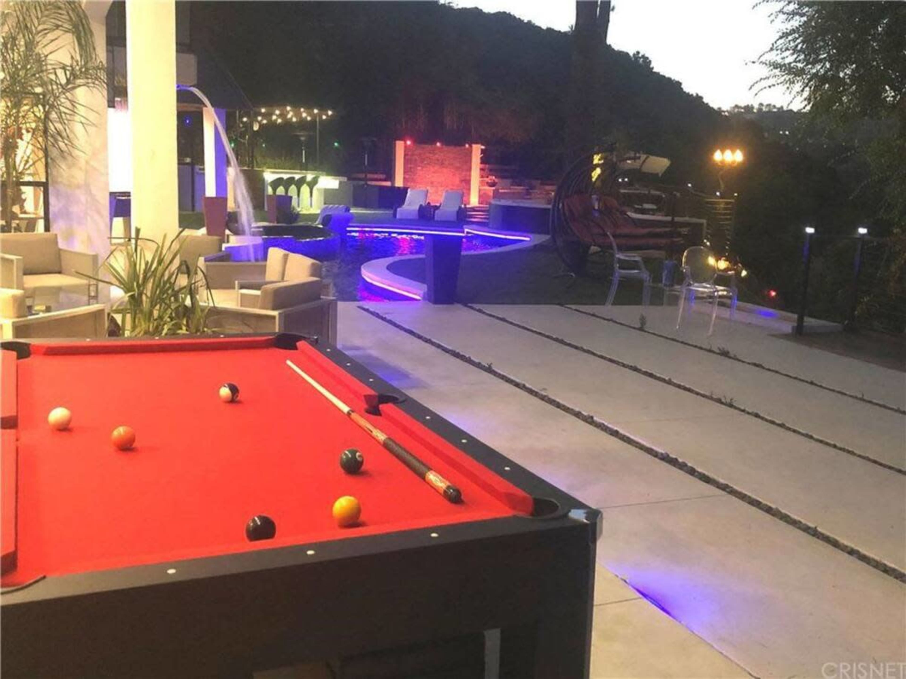 The scene shows a pool table with colorful balls in the foreground, and a well-lit outdoor area with a swimming pool and lounge seating in the background.