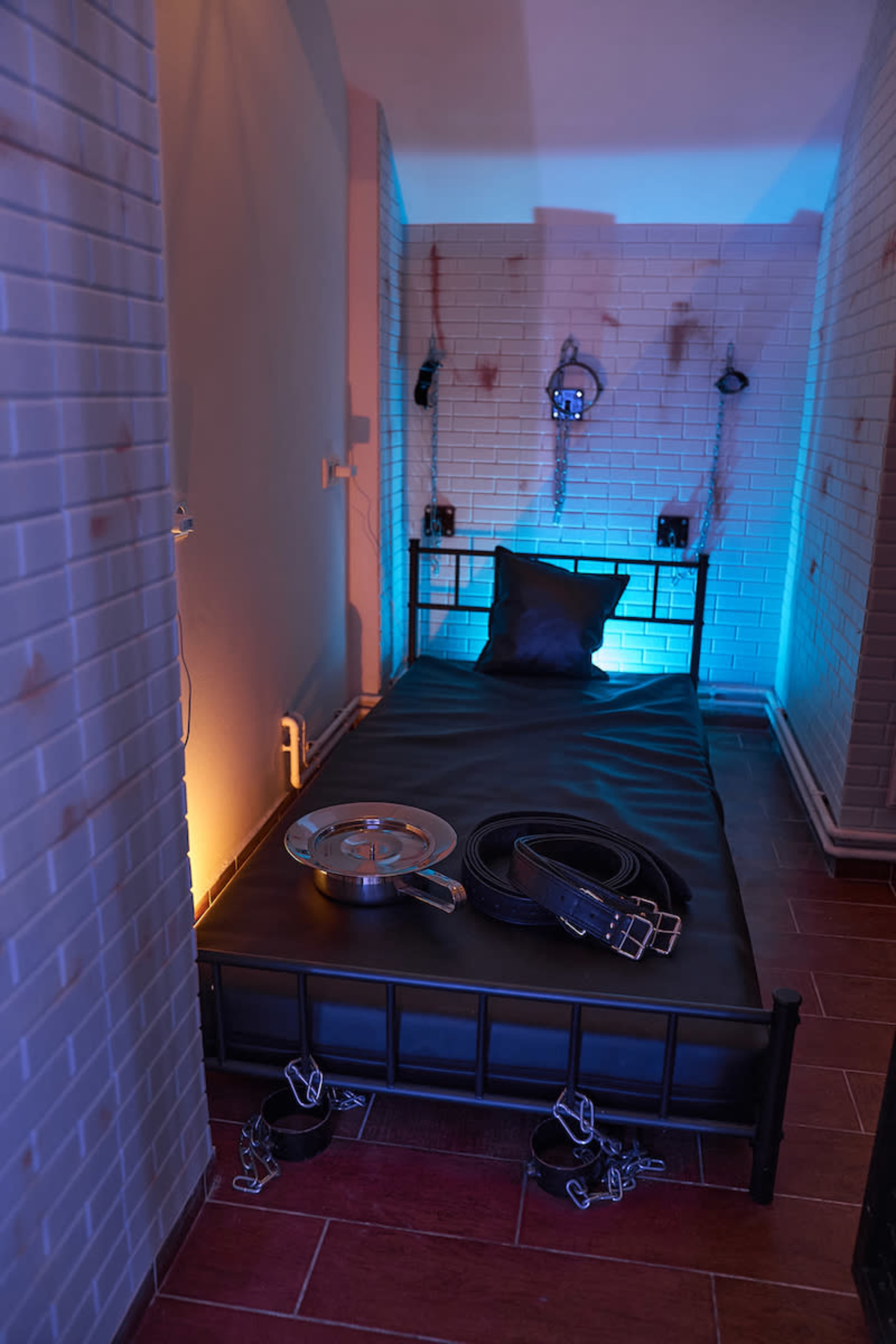 The image shows a minimalist room with a bed, restraints, and dim blue lighting against white brick walls.