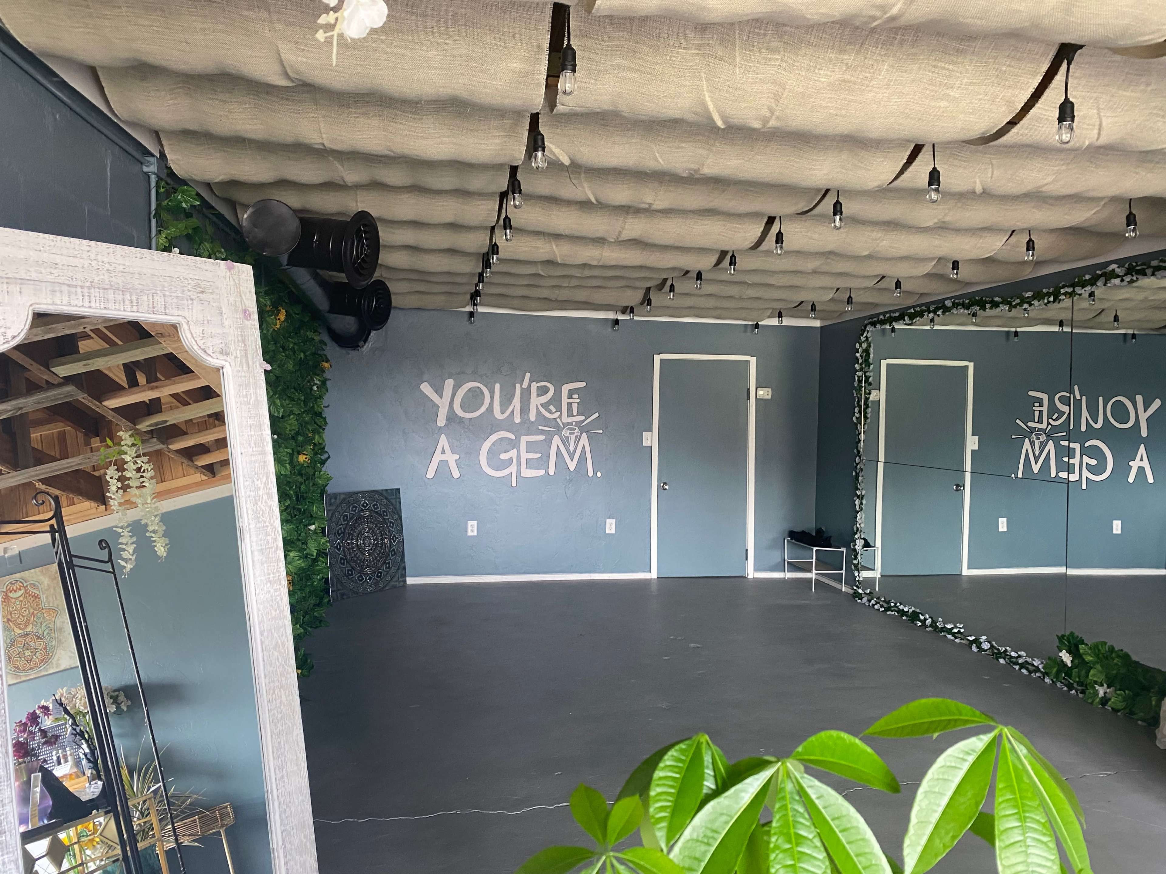 An empty room with a mirrored wall and a message on the wall that reads "YOU'RE A GEM," surrounded by greenery and soft lighting.
