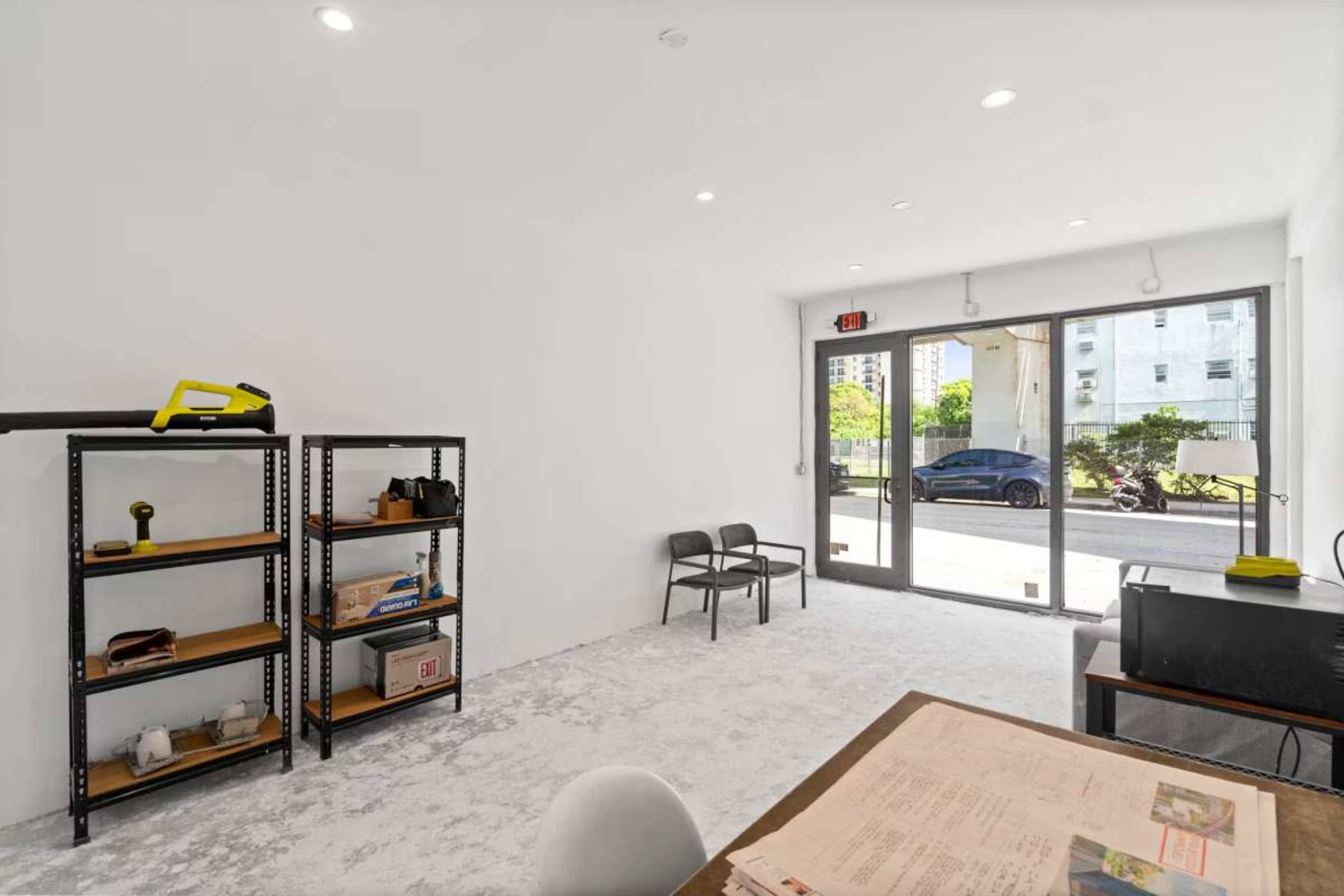 The image shows a minimalistic room featuring a large window, a table with documents, two chairs, and a bookshelf filled with various items.