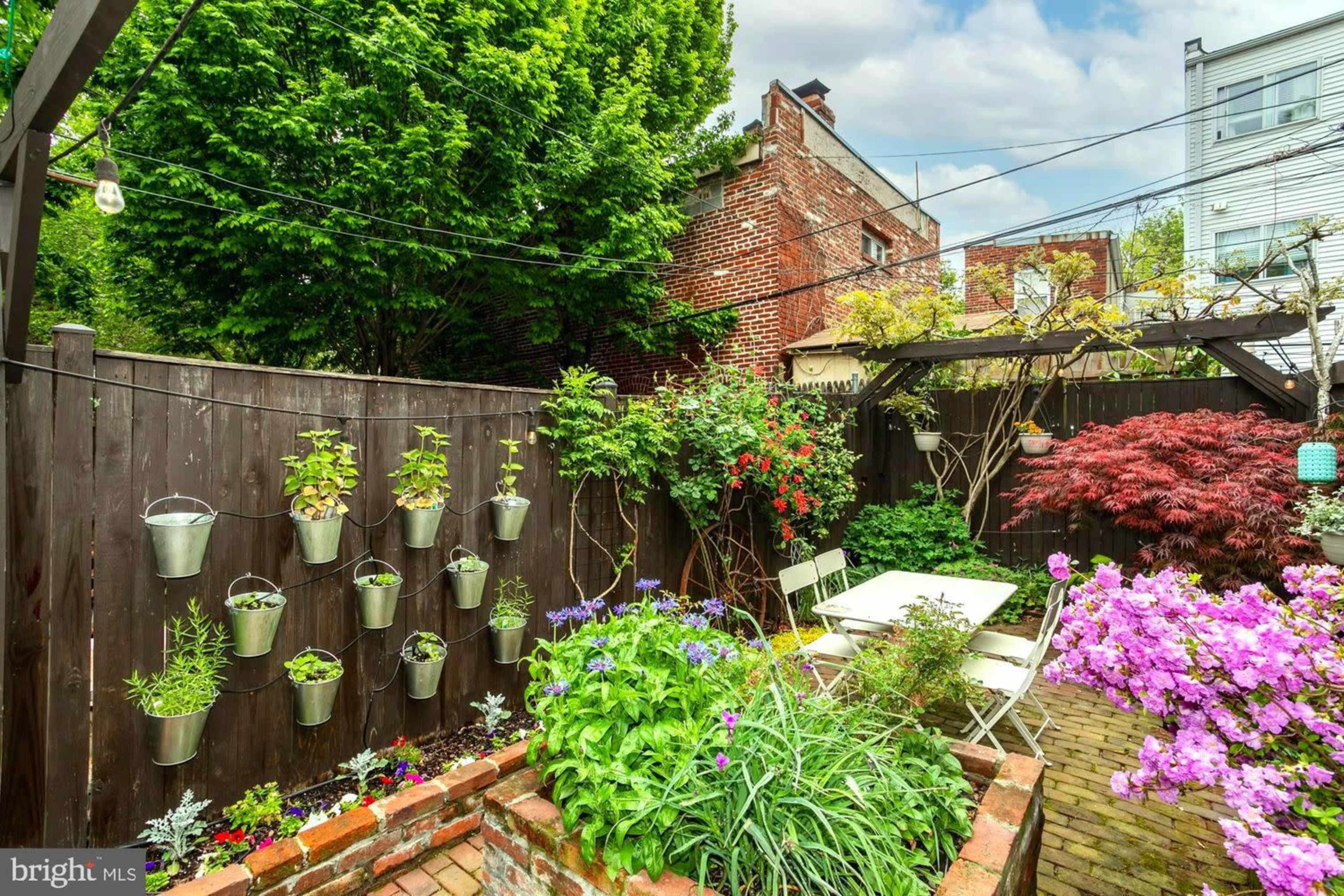 The image shows a small garden with hanging planters, colorful flowers, a brick pathway, and a seating area under a wooden arbor.